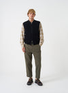 A male model wears the Universal Works Black Wool Fleece Zip Waistcoat zipped over a light shirt against a plain studio background. The shot highlights the regular fit, front zip closure, jersey collar, and angled front pockets.
