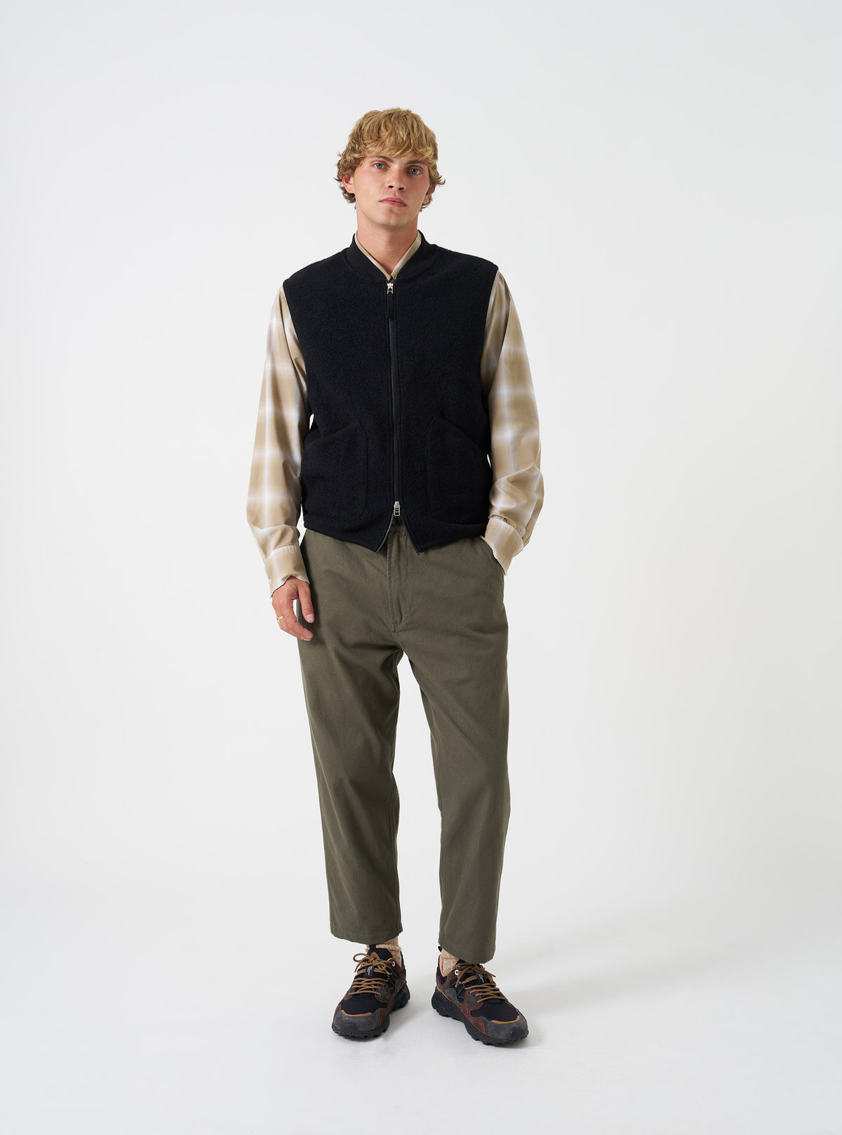 A male model wears the Universal Works Black Wool Fleece Zip Waistcoat zipped over a light shirt against a plain studio background. The shot highlights the regular fit, front zip closure, jersey collar, and angled front pockets.