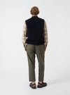 A male model showcases the Universal Works Black Wool Fleece Zip Waistcoat worn unzipped over layers against a neutral studio background. The image highlights the relaxed silhouette, open front styling, and textured fleece material.