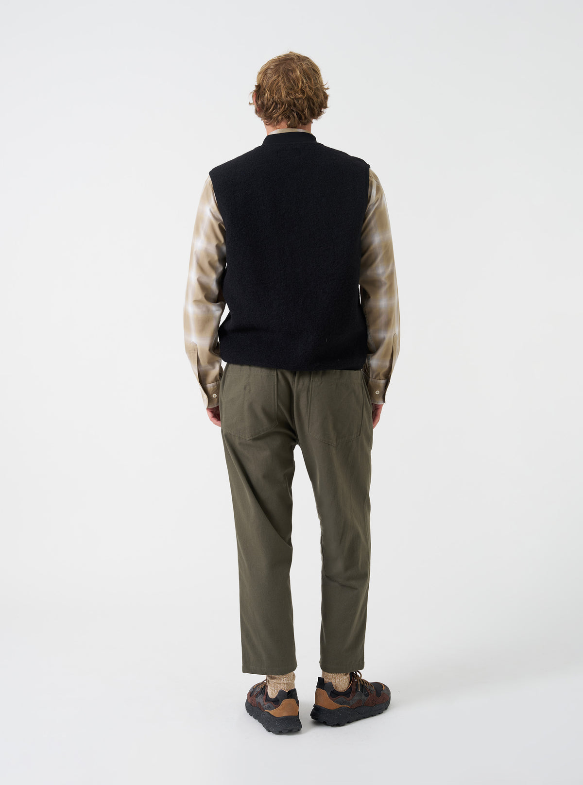 A male model showcases the Universal Works Black Wool Fleece Zip Waistcoat worn unzipped over layers against a neutral studio background. The image highlights the relaxed silhouette, open front styling, and textured fleece material.