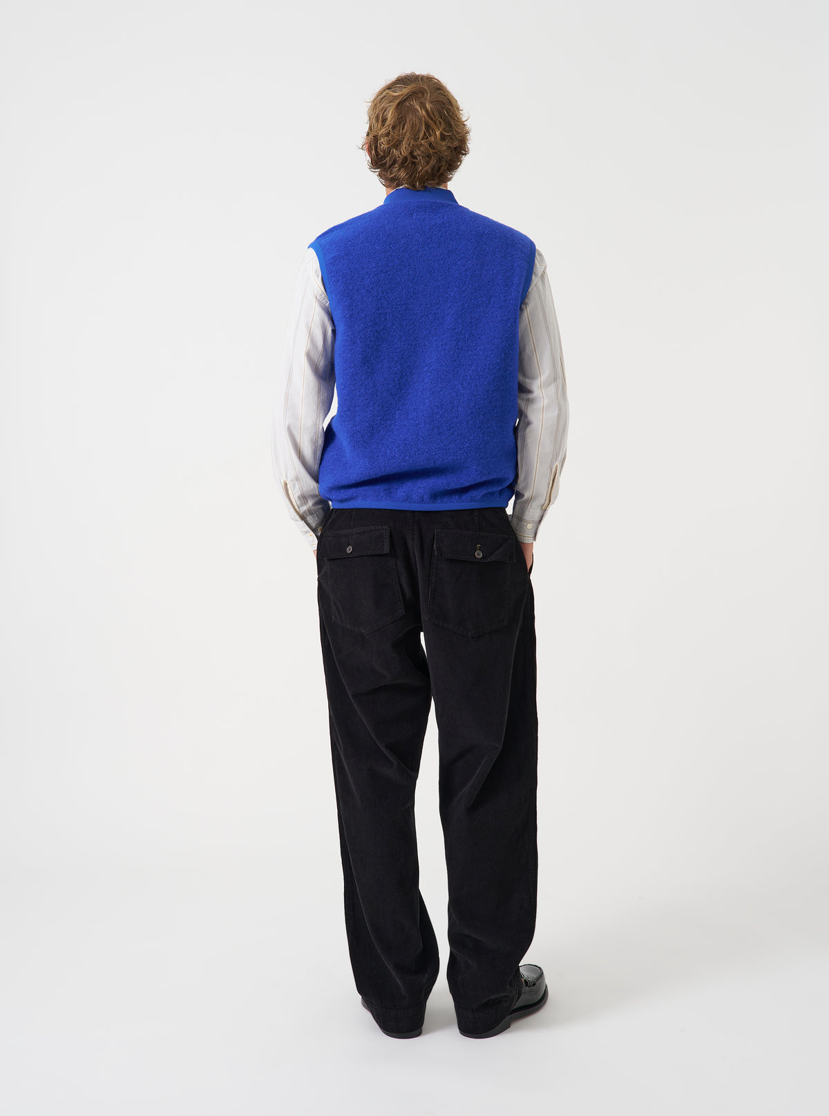 A male model showcases the Universal Works Blue Wool Fleece Zip Waistcoat worn unzipped over layers against a neutral studio background. The image highlights the open-front styling, relaxed fit, and cozy wool fleece material.