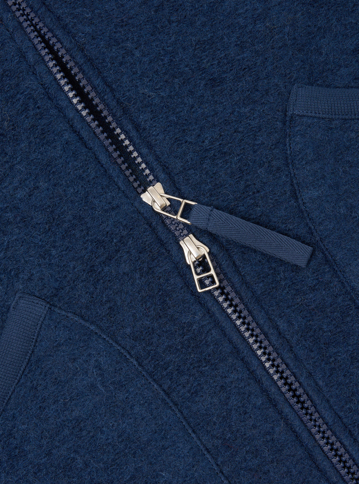Close-up detail shot of the Indigo Wool Fleece Zip Waistcoat on a plain background. Focuses on the metallic front zipper hardware, rounded collar edge, and rich indigo blue fleece texture of the garment.