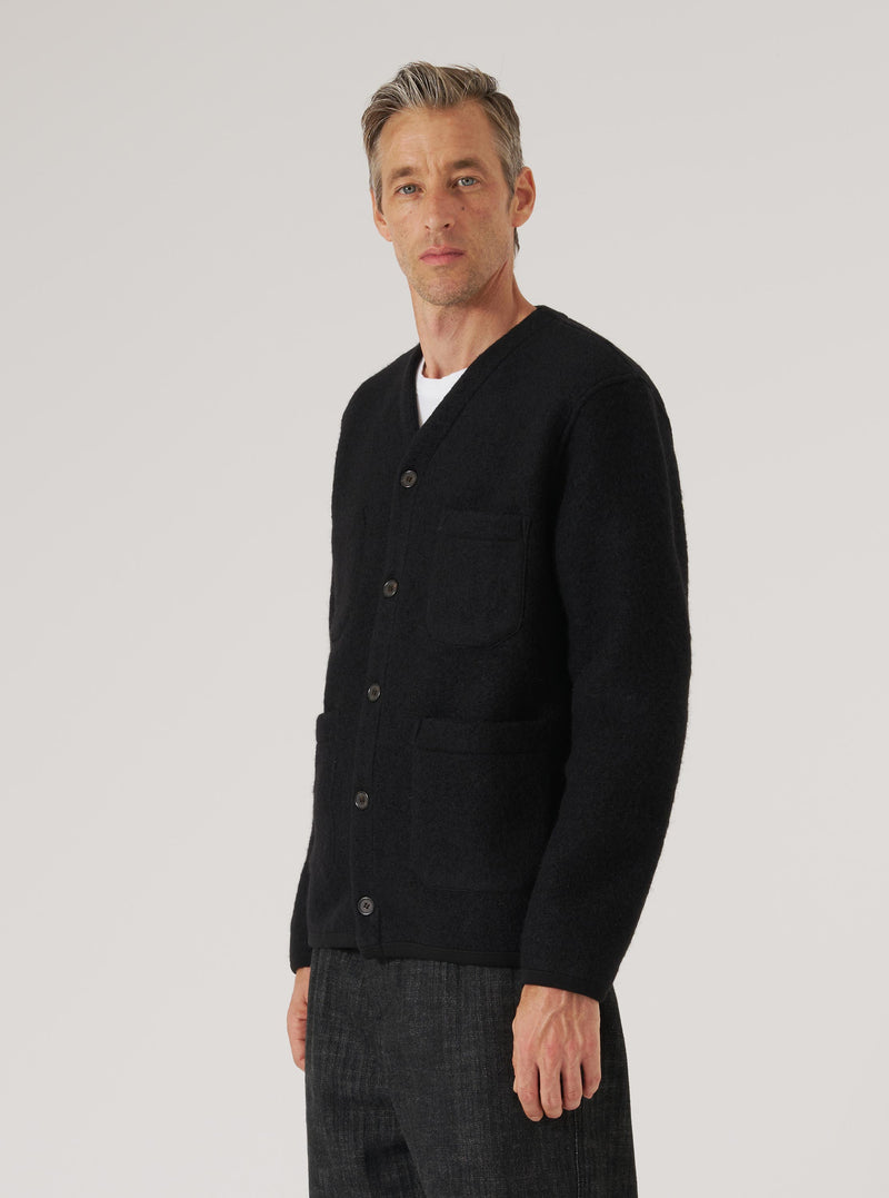 A man models a Black Wool Fleece Cardigan in front of a plain background. The cardigan has a V-neck and button closure, with patch pockets. He wears a white t-shirt underneath.