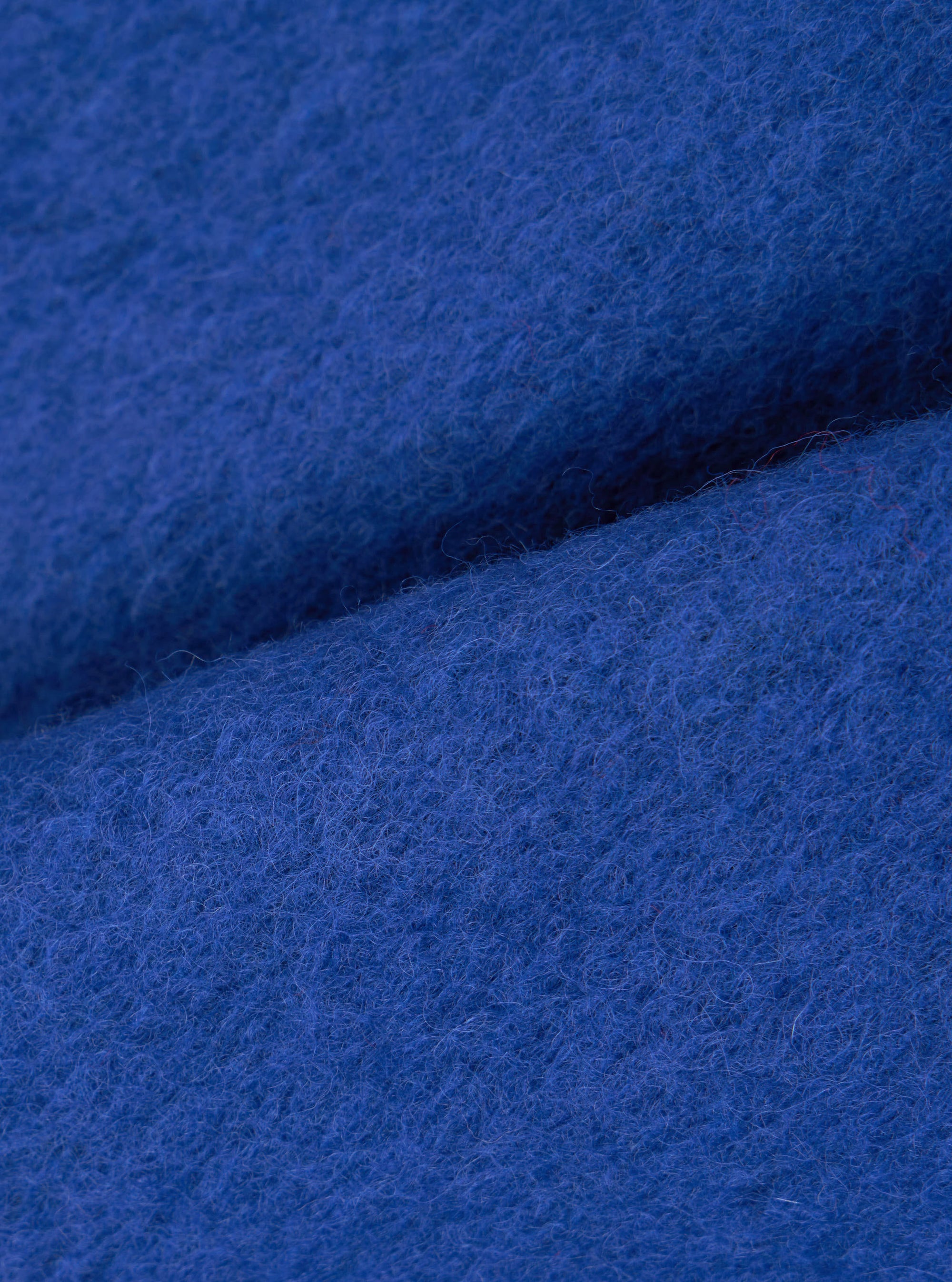 Close-up fabric detail of the blue wool fleece cardigan, showing the dense, brushed texture of the cobalt blue wool. Fine fibres are visible across the surface, with soft folds creating depth and shadow across the frame.