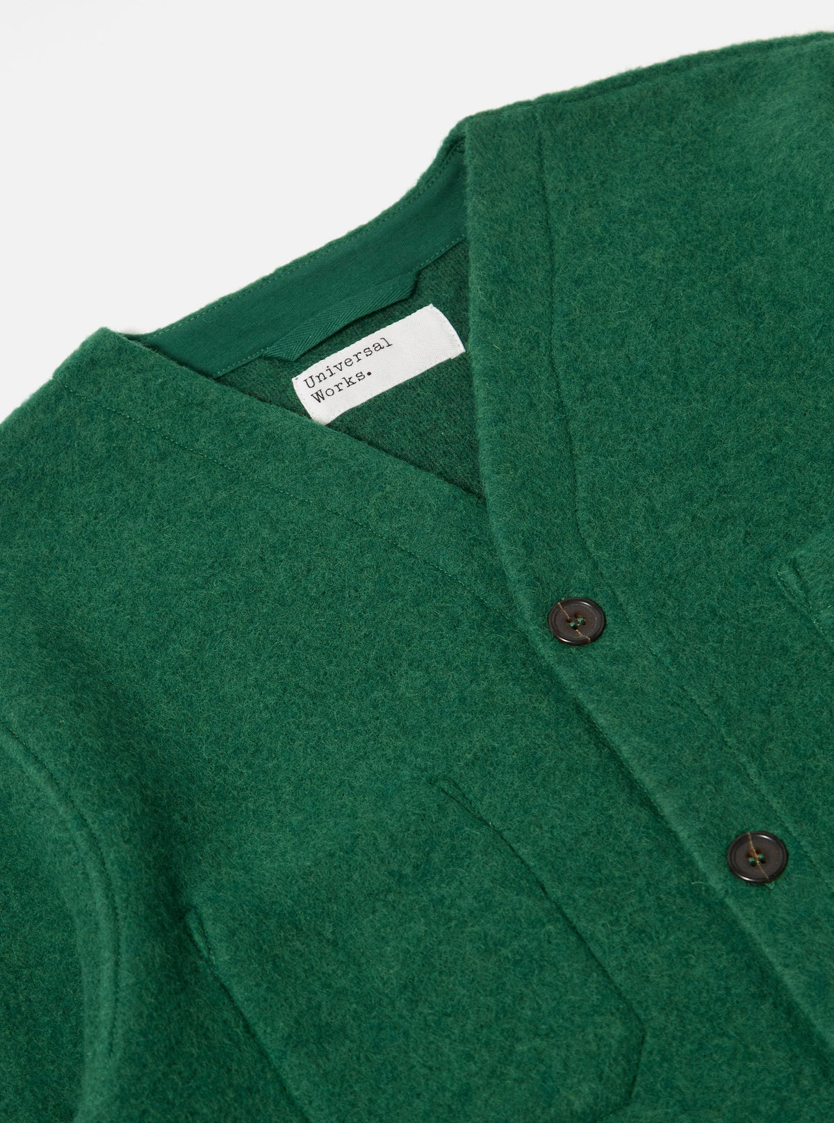 Close-up of a Green Wool Fleece Cardigan on a white surface. The cardigan has brown buttons and a visible tag. The wool has a textured, slightly fuzzy appearance.
