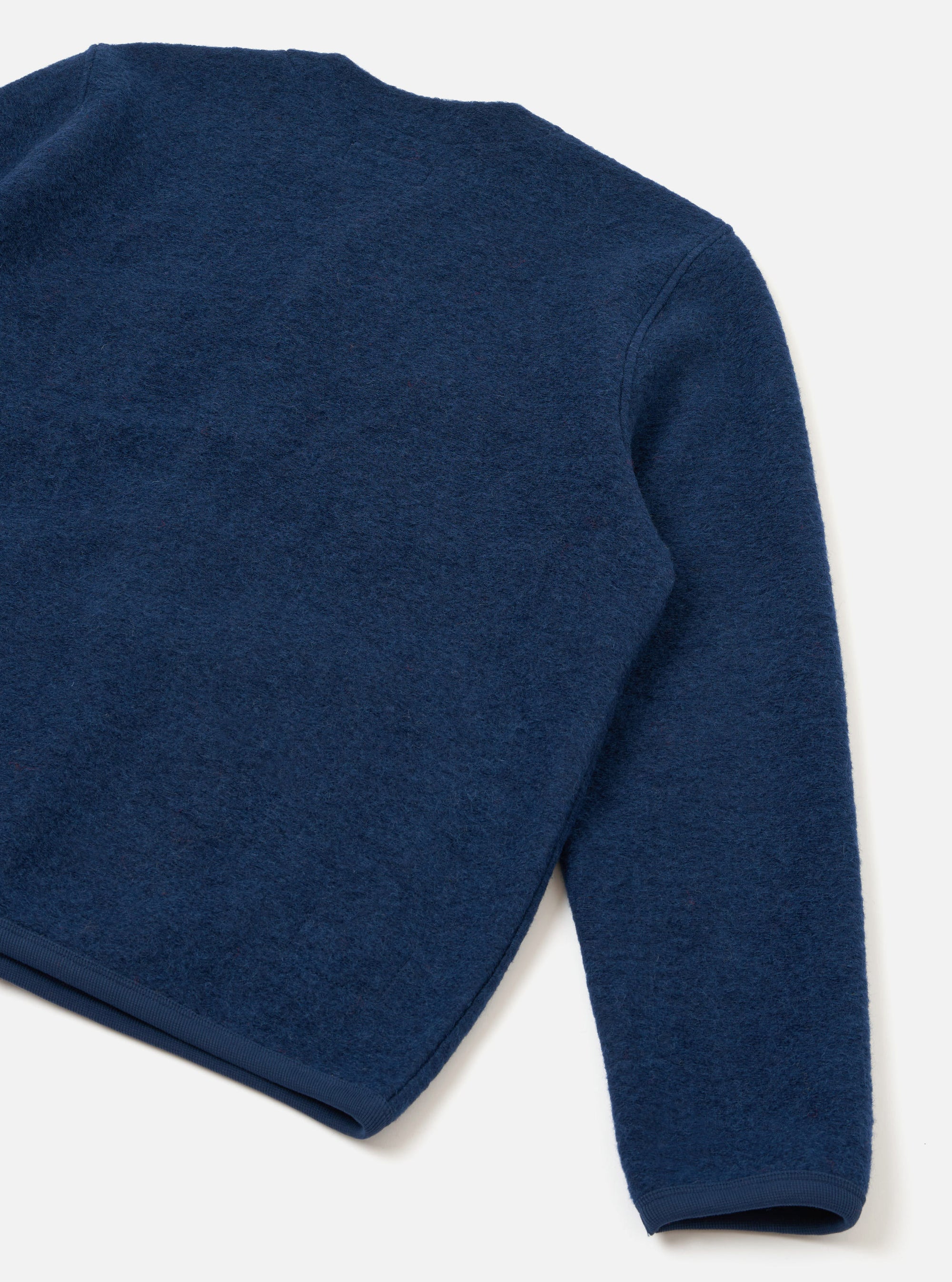 Close-up flat lay of an indigo wool fleece cardigan on a white studio surface, showing the back panel and one sleeve. The boiled wool texture is clearly visible across the body. A ribbed hem and ribbed cuff are visible at the bottom edges.