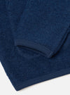 Close-up detail of an indigo wool fleece cardigan laid flat on a white surface. The shot focuses on the sleeve cuff and lower hem, both finished with ribbed knit trim in matching indigo. The boiled wool fleece body shows a dense, textured surface with visible fibre variation throughout.