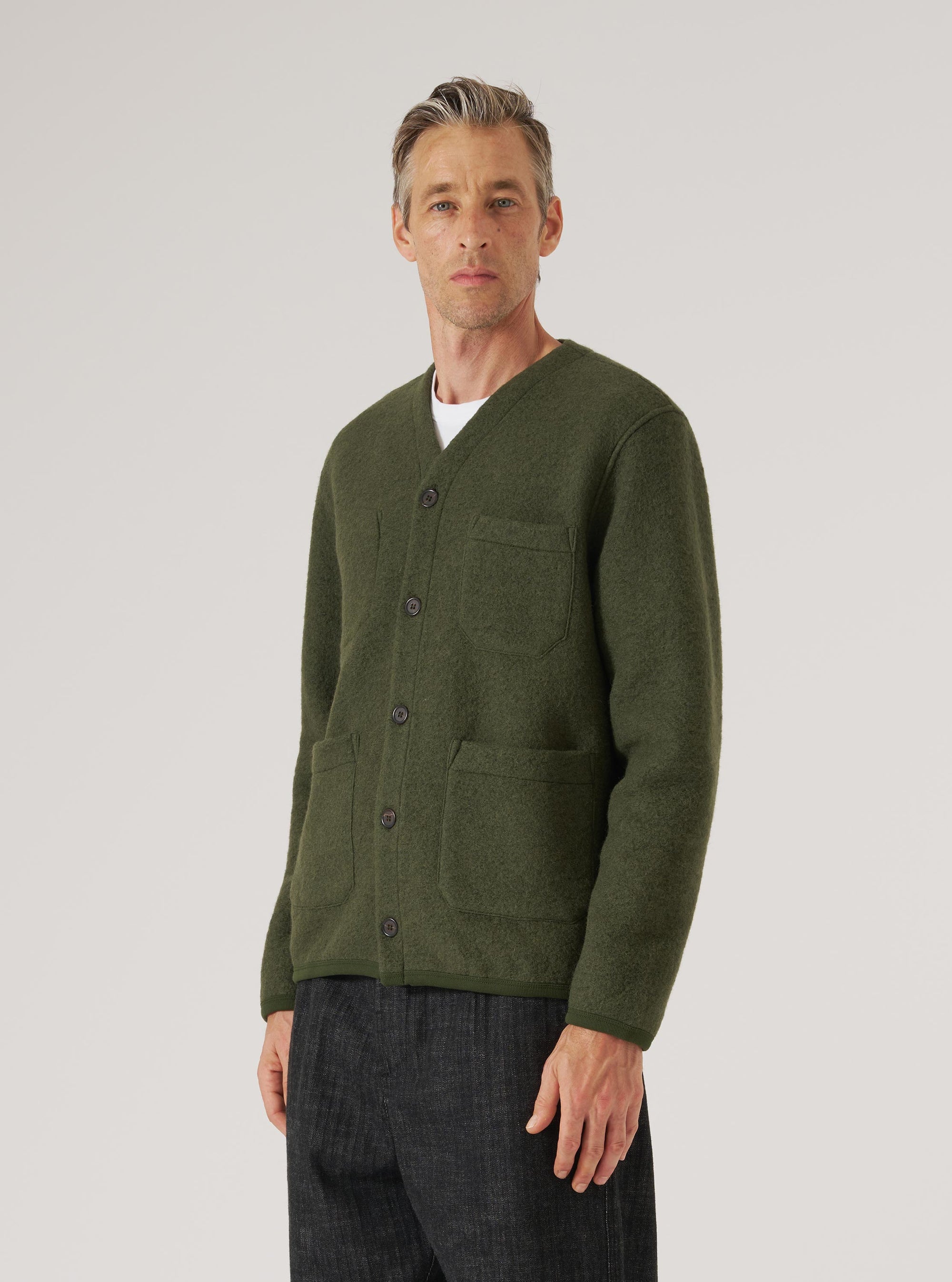 Male model wearing an olive wool fleece cardigan, buttoned, over a white crew-neck tee, with dark trousers. V-neck, four dark buttons, one chest patch pocket and two lower patch pockets. Textured boiled wool fabric. Shot from a slight angle, cropped at the thigh, on a light grey studio backdrop.