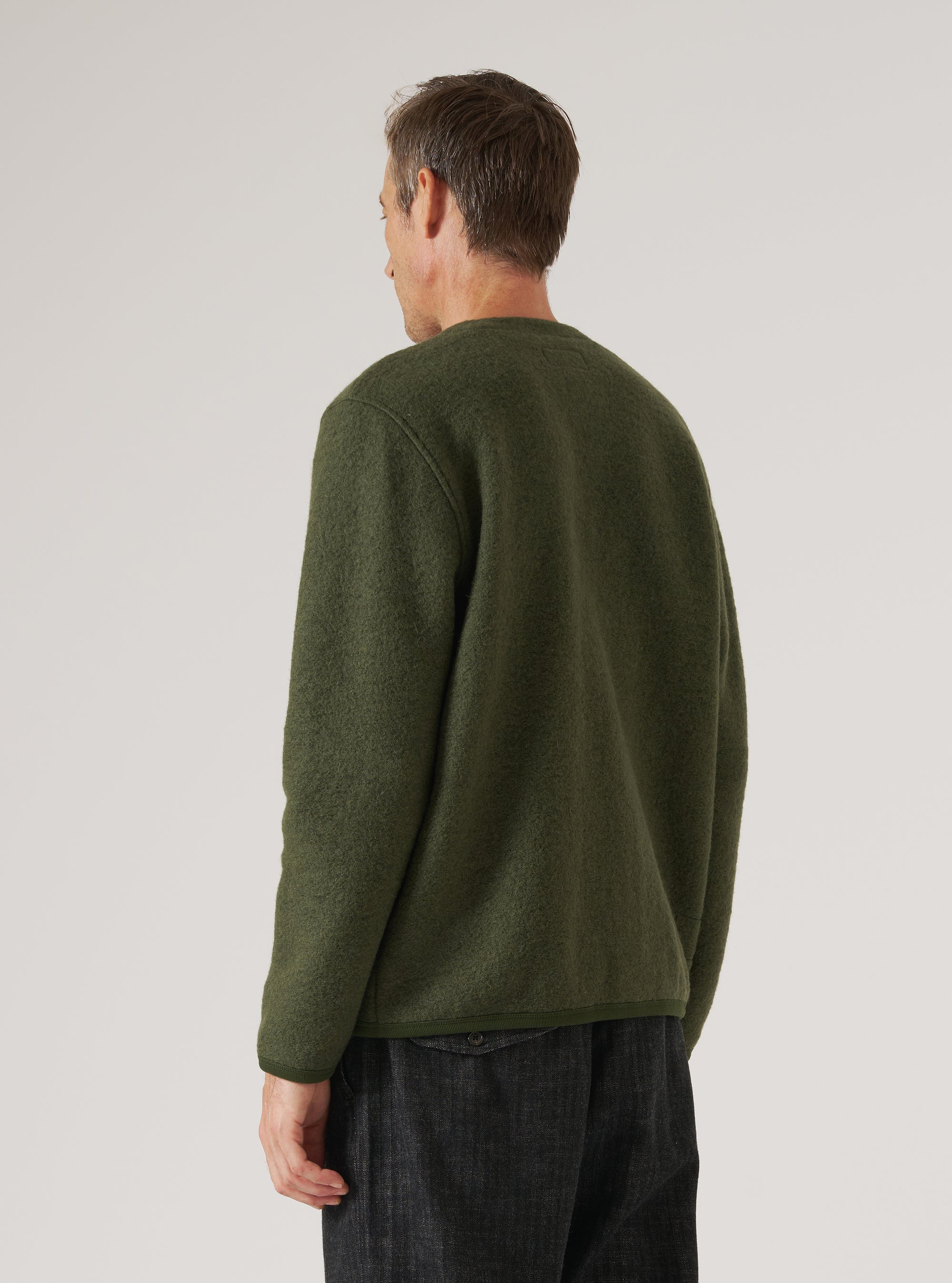 Back view of a male model wearing an olive wool fleece cardigan on a light grey studio backdrop. The textured, brushed fleece fabric is shown clearly across the back and full-length sleeves. A crew neckline and ribbed hem are visible. The cardigan is worn untucked over dark charcoal trousers. Shot cropped from mid-thigh up.