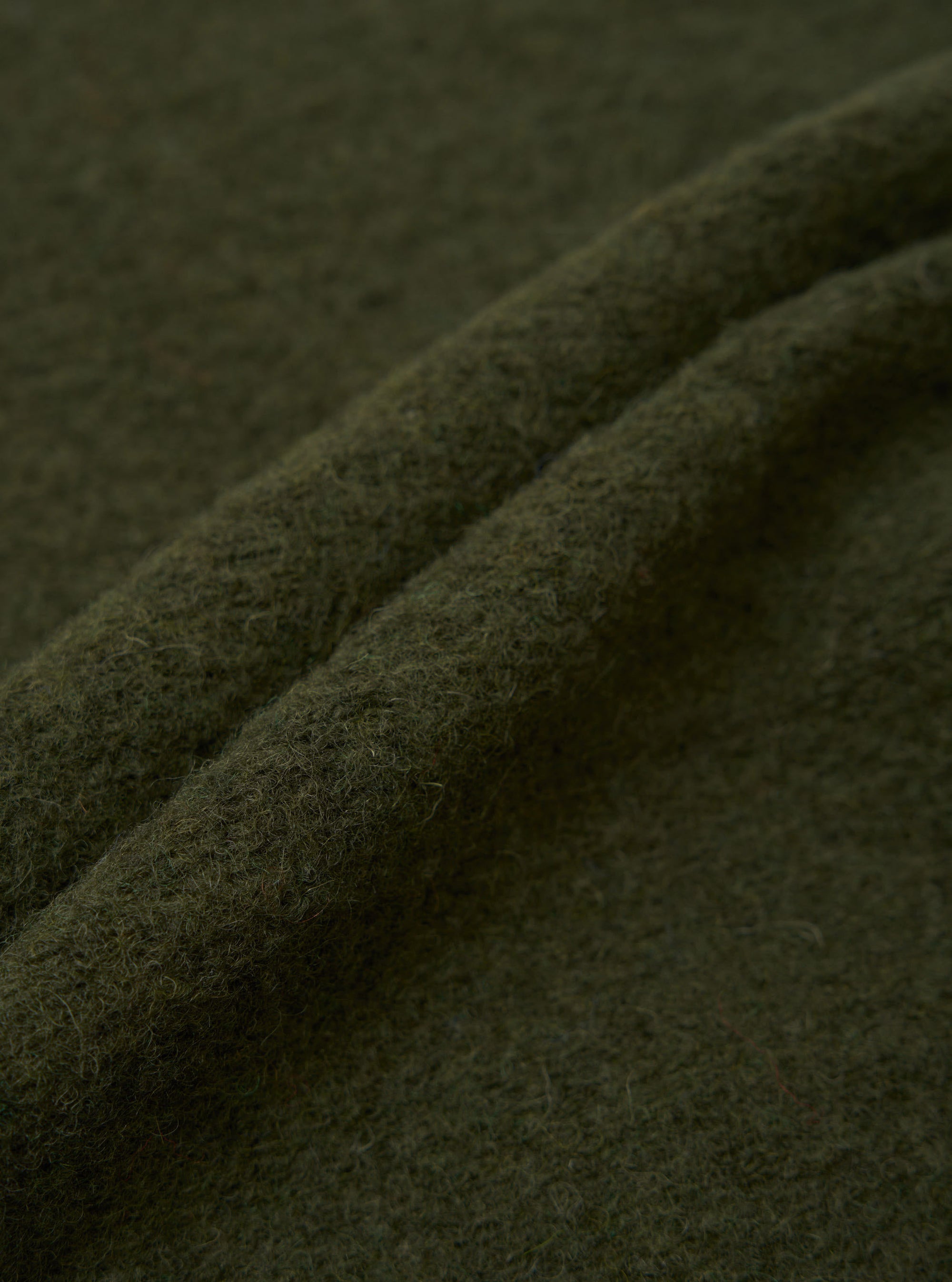 Close-up of olive wool fleece fabric, showing soft, lightly brushed texture with fine surface fibres and natural drape. The deep khaki green cloth is folded to reveal its thickness and fluffy handle. No garment details or construction are visible in this fabric detail shot.