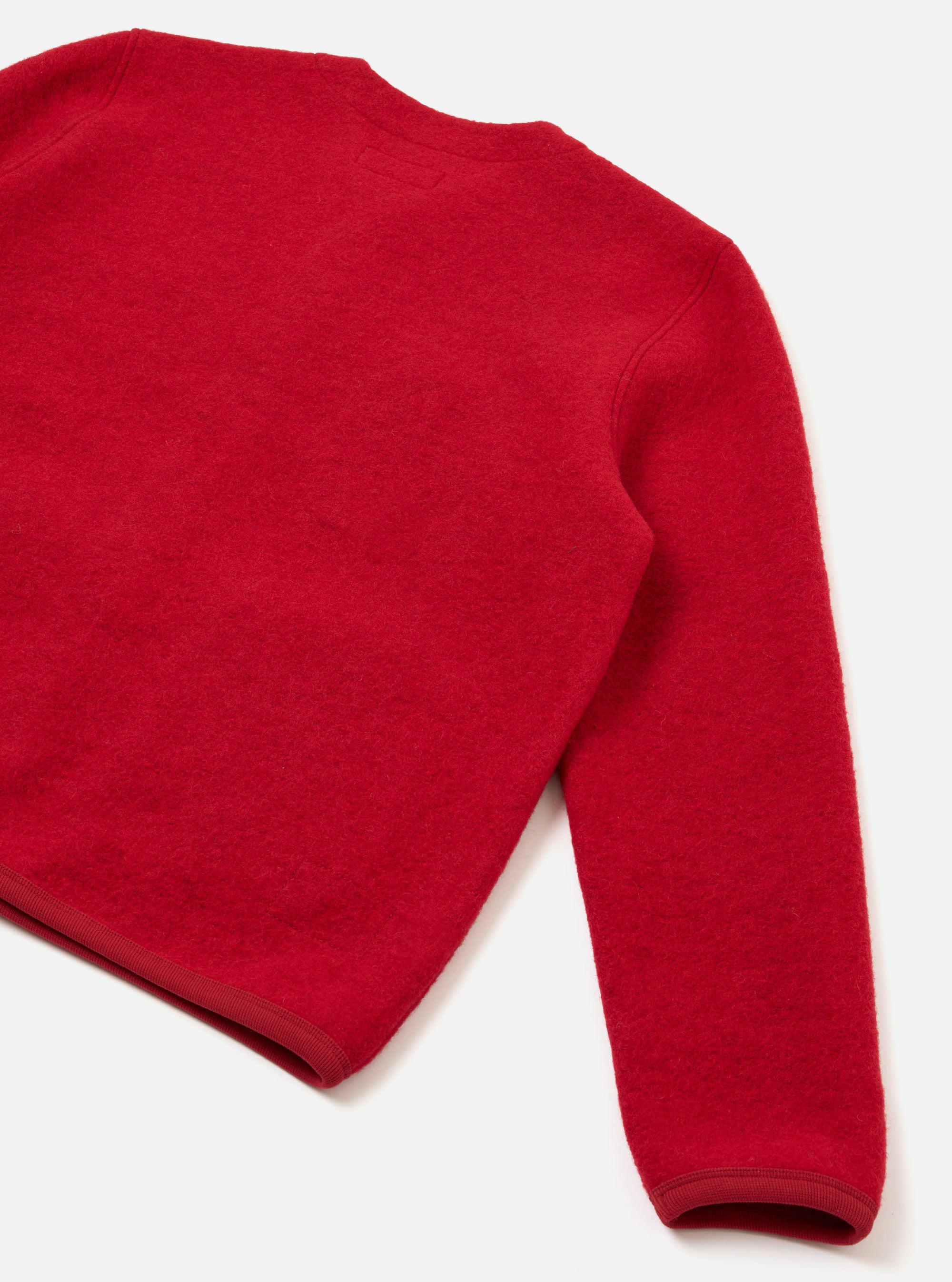 Close-up flat lay of a scarlet wool fleece cardigan on a white surface, showing the back panel and one full sleeve. The boiled wool texture is visible across the body. A narrow ribbed grosgrain hem band and matching cuff trim finish the edges in the same scarlet tone.