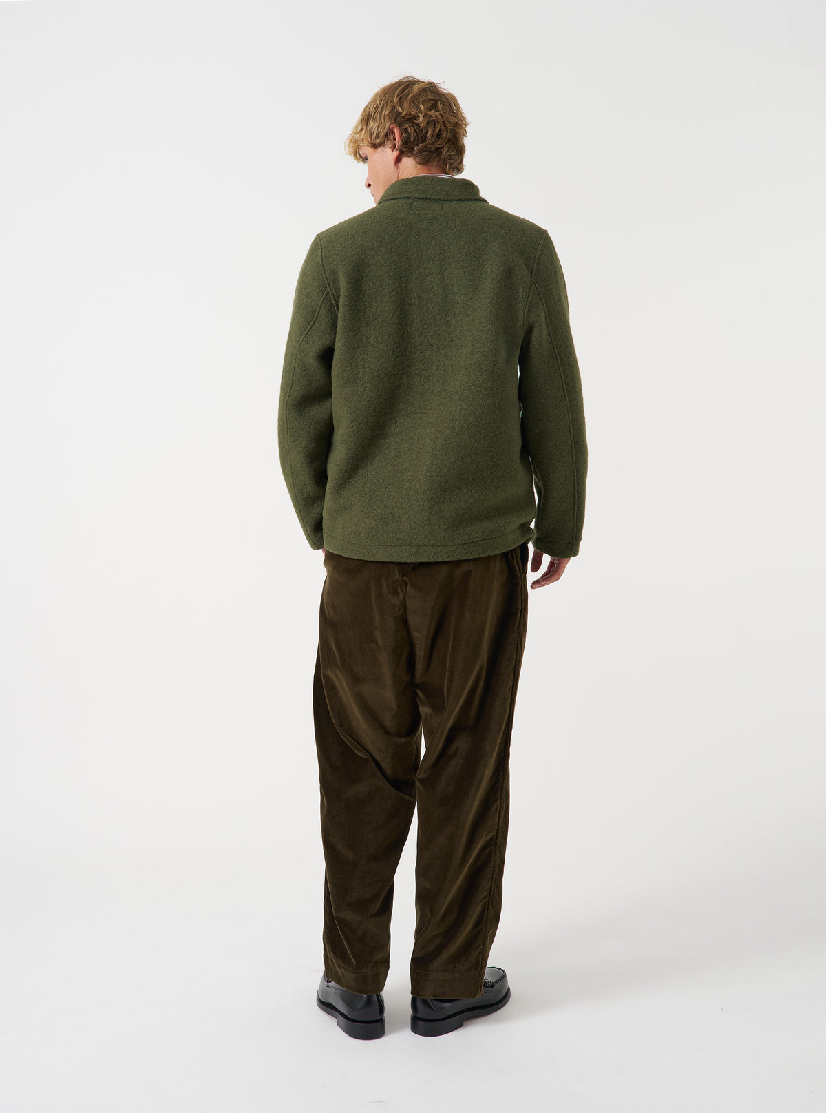 Angled view of a model wearing the Olive Wool Fleece Field Jacket in a studio setting. The model turns slightly, showing the fit, sleeve length, and rich olive green wool fleece material from a side perspective.
