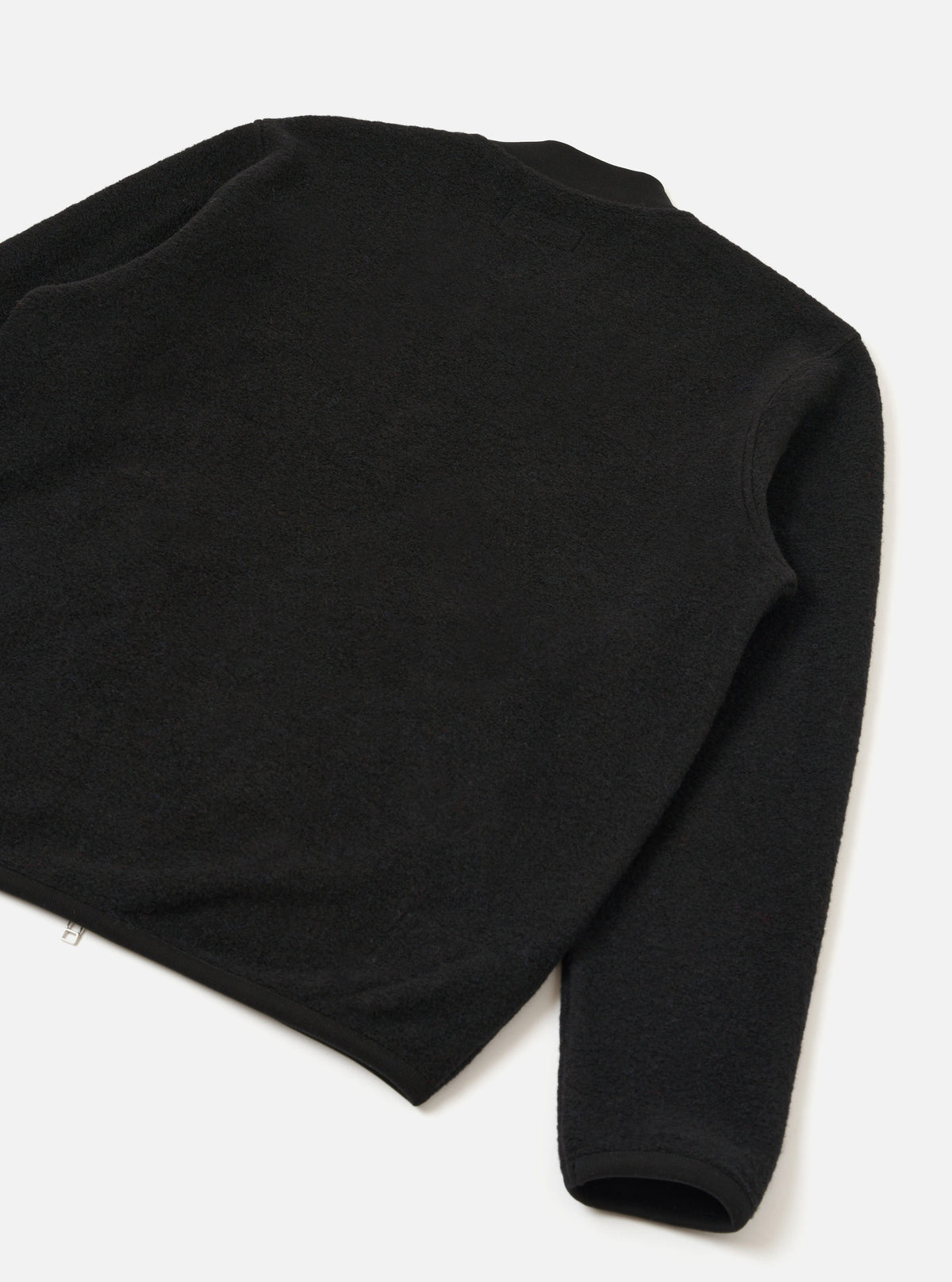 Black wool fleece zip bomber shot as a flat lay on a white surface, angled to show the back and one sleeve. The textured fleece is visible across the body and sleeve. A ribbed cuff is visible at the hem of the sleeve. A small silver zip pull is visible at the lower left hem.