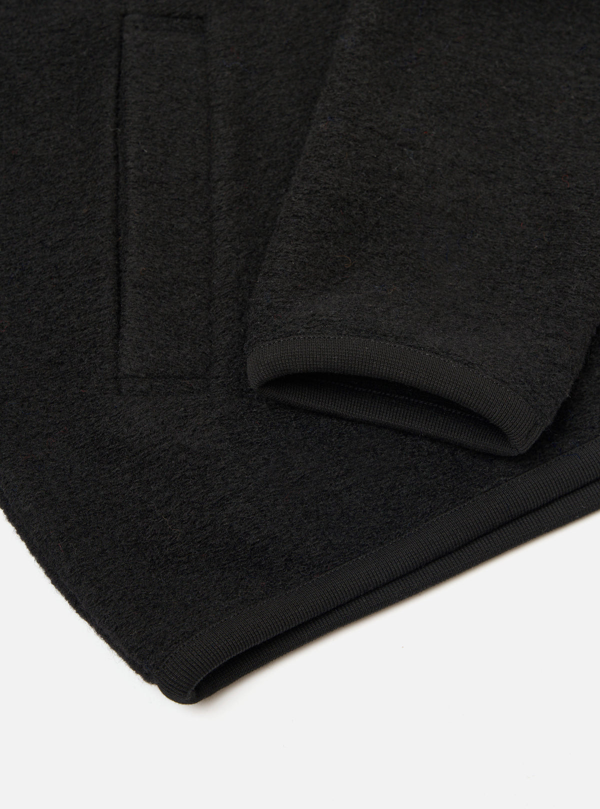 Close-up flat lay of a black wool fleece zip bomber on a white surface. The shot focuses on the lower sleeve and hem, showing the ribbed knit cuff and ribbed hem band. The dense, textured wool fleece fabric is visible in detail across the body and sleeve.