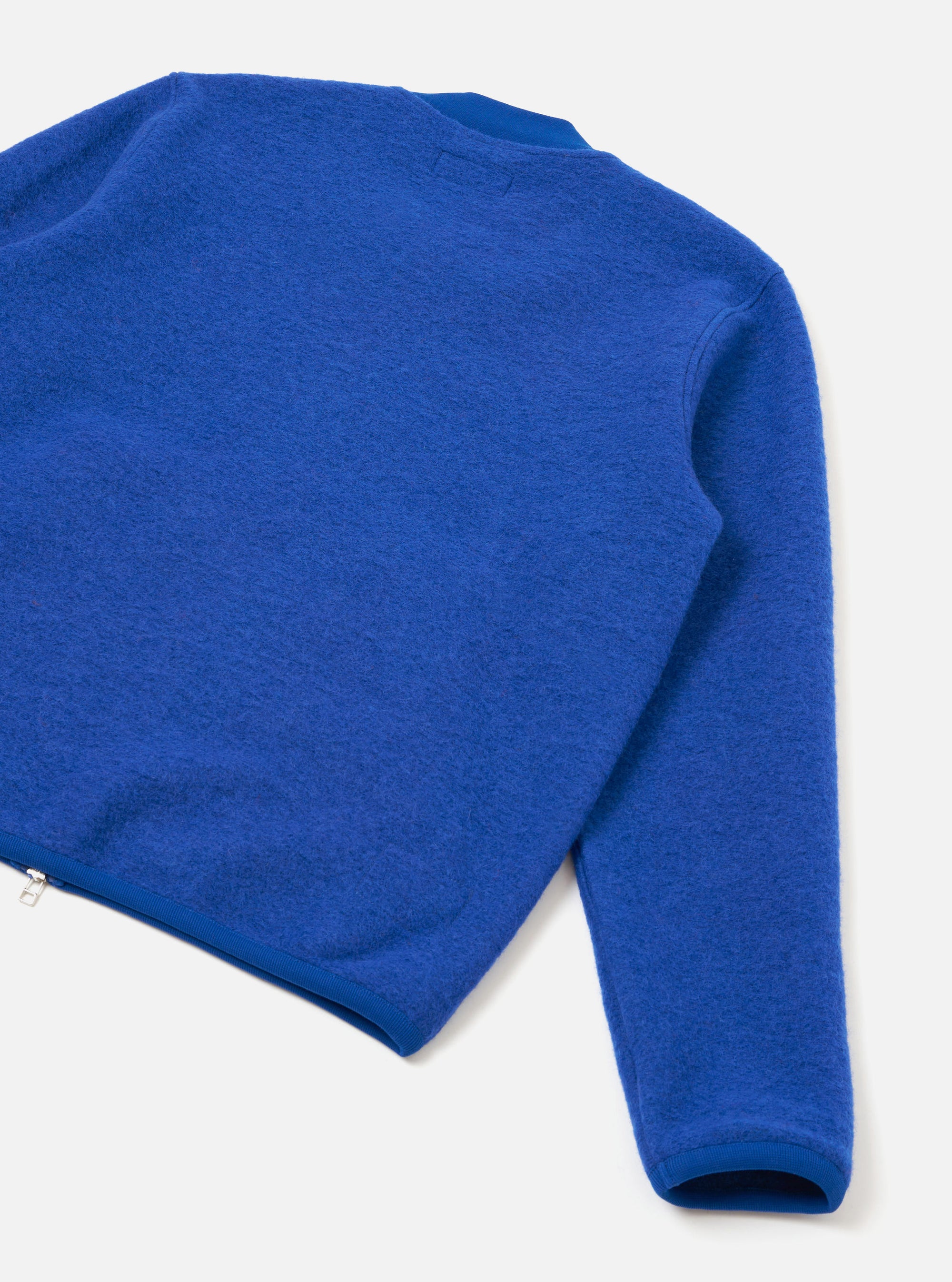 Blue wool fleece zip bomber laid flat on a light grey surface, shot from above at an angle. The image shows the back and one sleeve, highlighting the brushed wool fleece texture. A silver zip is visible at the lower left hem. Ribbed cuffs and hem trim contrast with the fluffy fleece body.