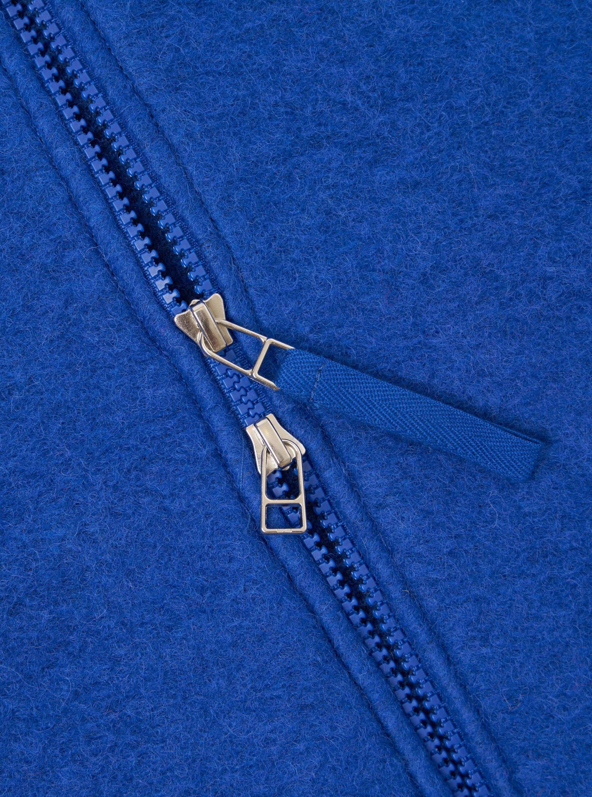Close-up detail of the double zip fastening on a blue wool fleece zip bomber. Two gold-tone metal zip sliders sit on blue plastic teeth, with a woven blue zip pull tab. The brushed wool fleece texture is visible across the royal blue fabric.