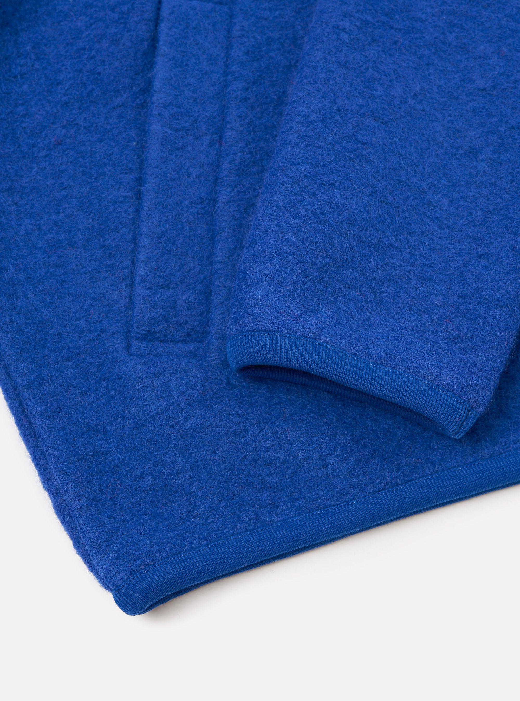 Close-up flat lay of a blue wool fleece zip bomber on a white surface. The textured boiled wool fabric is shown in detail alongside a ribbed knit cuff at the sleeve end and a matching ribbed hem. A front zip placket is visible at the top left of the frame.