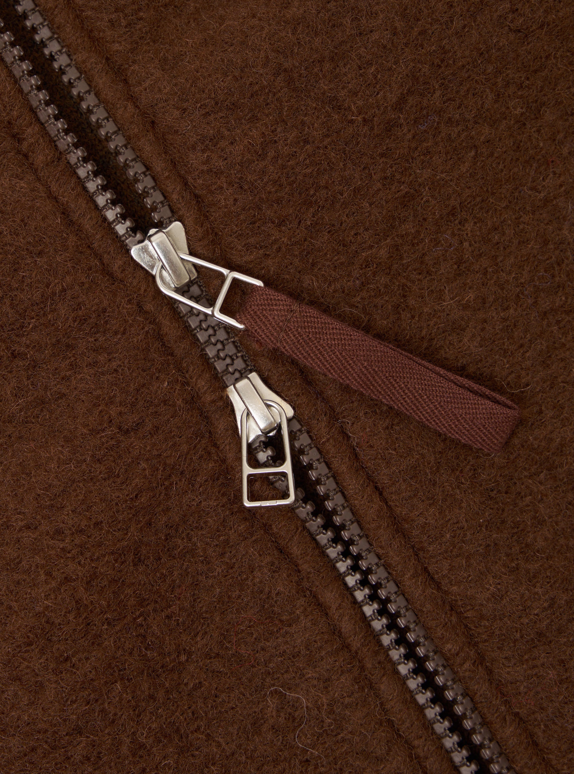 Close-up detail of the double zip on a cacao wool fleece bomber. Two gunmetal zip sliders with silver hardware sit mid-track on a dark resin chain. A woven fabric zip pull in deep burgundy-brown is attached to the upper slider. The brushed wool fleece ground shows a dense, hairy texture throughout.