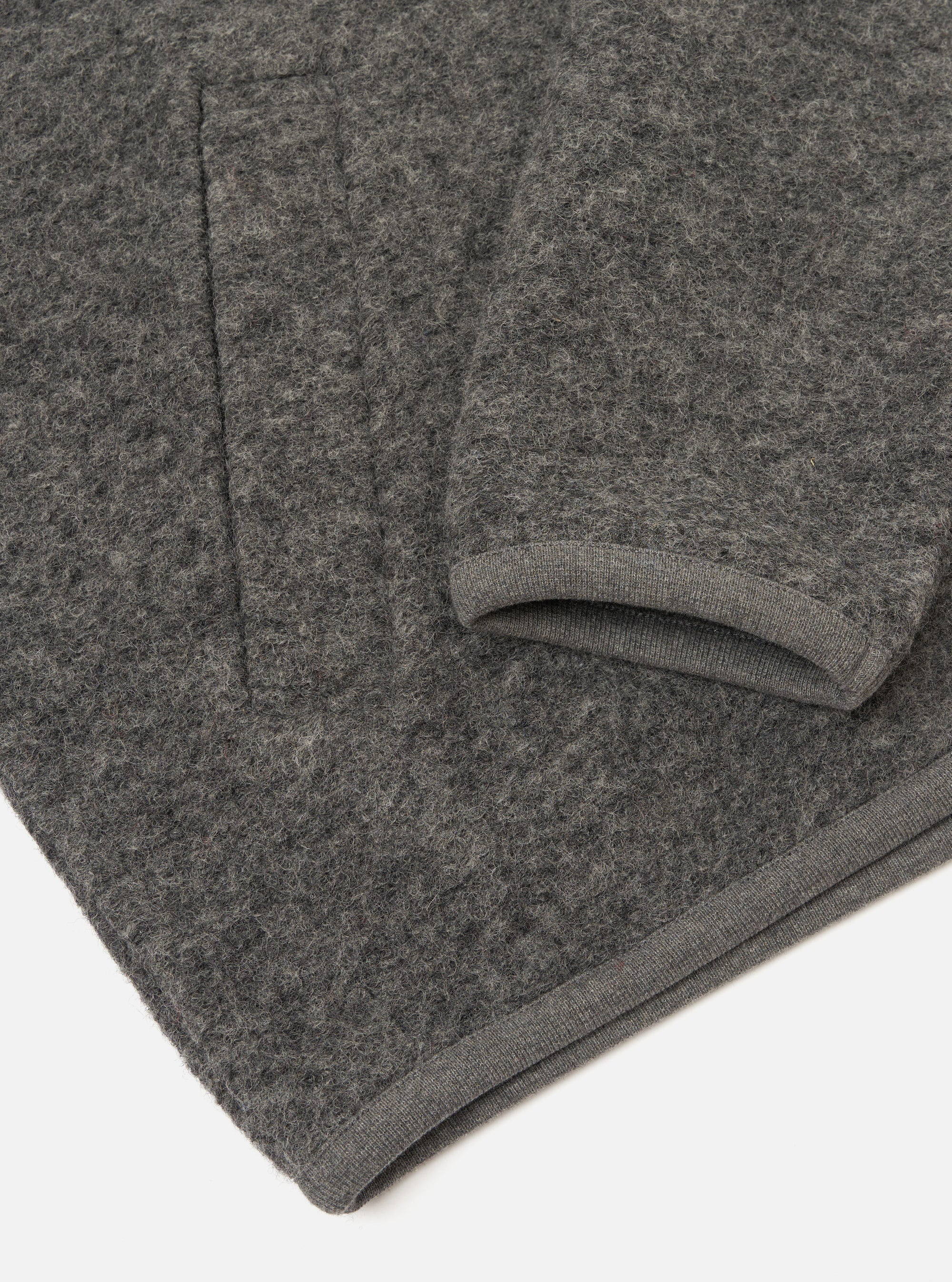Close-up flat lay of a grey marl wool fleece zip bomber, folded to show the sleeve and hem. The boiled wool fleece has a dense, nubby texture throughout. A ribbed knit cuff is visible at the sleeve end, and a matching ribbed hem strip runs along the bottom edge. Shot on a white studio surface.