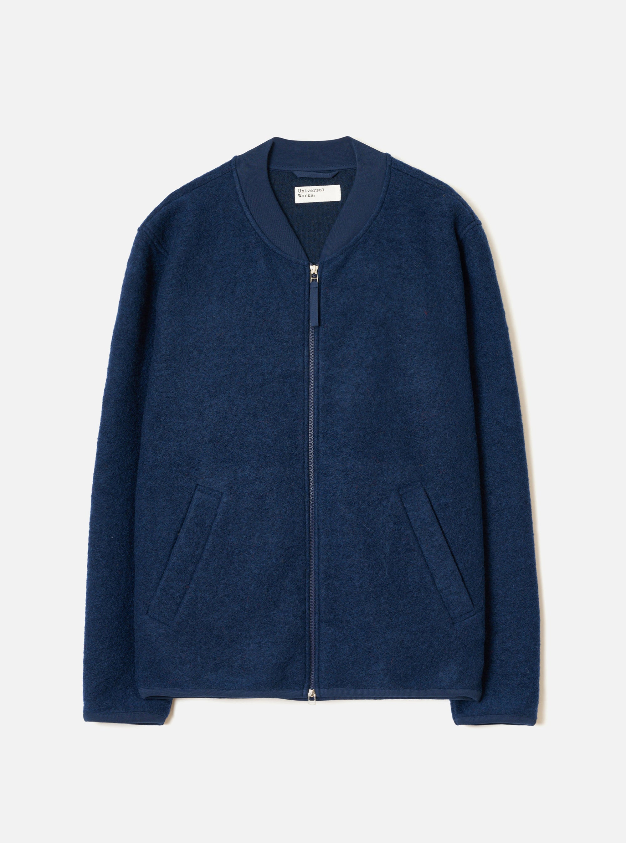 Indigo wool fleece zip bomber laid flat on a light grey studio backdrop. The jacket features a ribbed bomber collar in matching navy fabric, a full-length silver zip, two welt hip pockets, and ribbed cuffs. The boiled wool fleece has a dense, textured surface. A Universal Works woven label is visible at the neck.