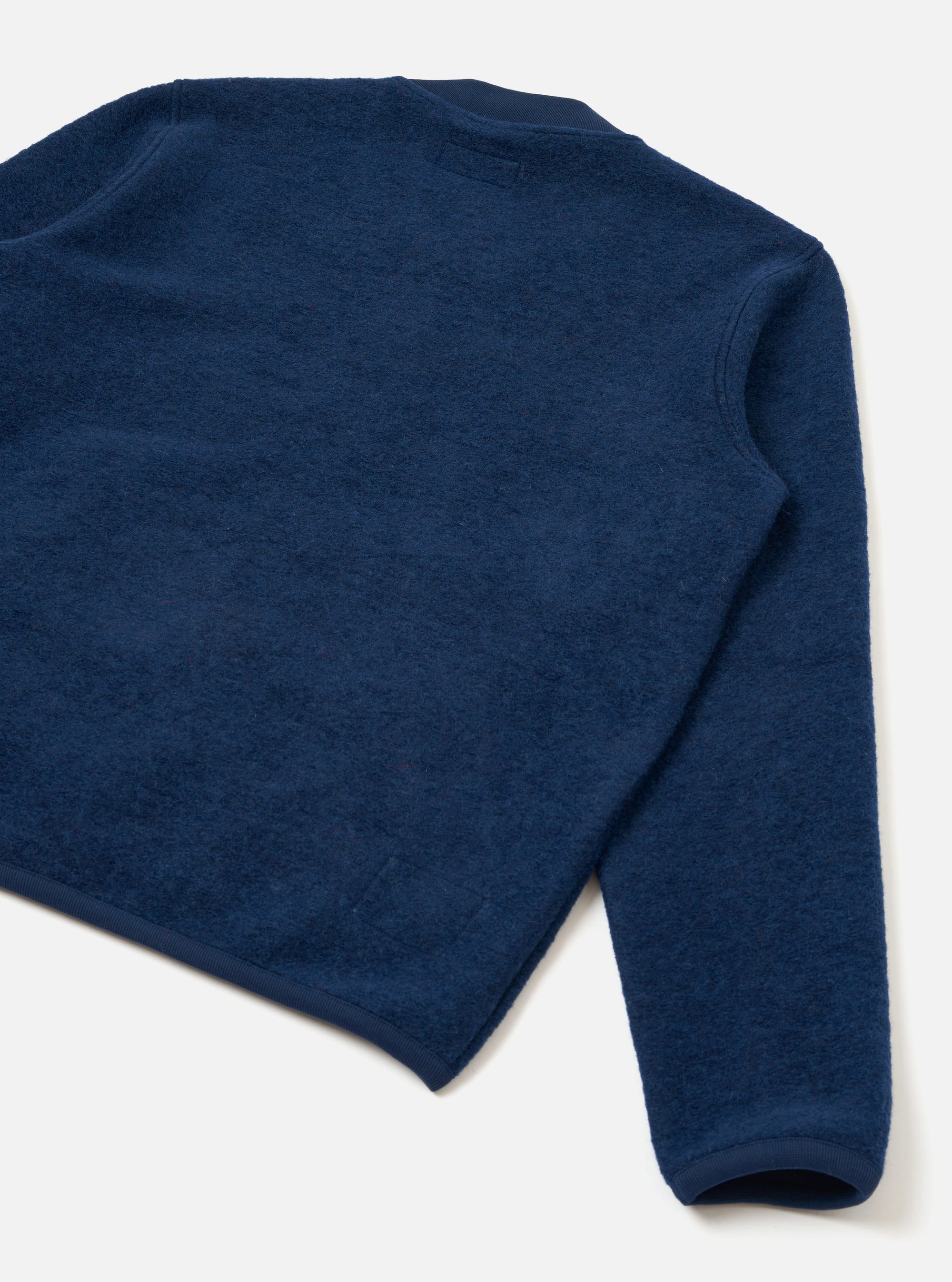 Flat lay of an indigo wool fleece zip bomber on a white studio surface, shot from above at an angle. The back and one sleeve are visible, showing a dense, brushed wool fleece texture. Navy bound seams run along the armhole and hem. The cuff is finished with a matching navy ribbed band.