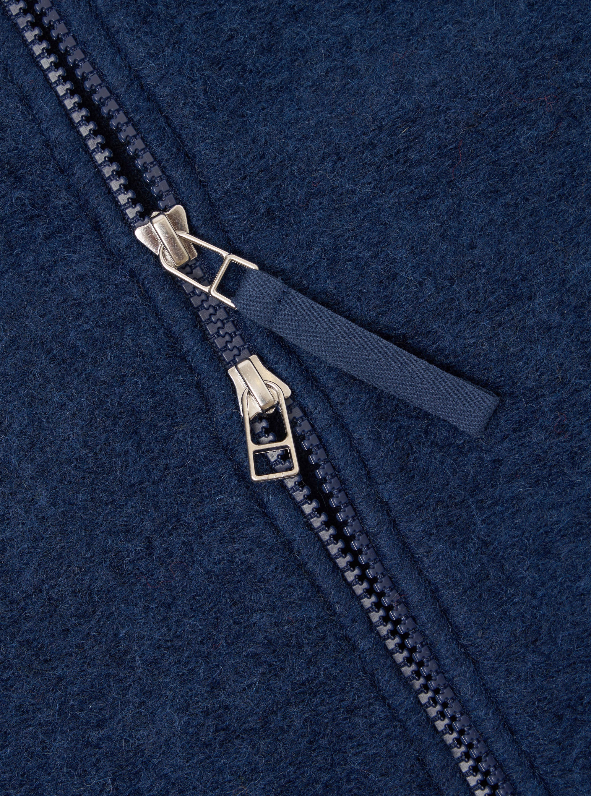 Close-up detail of the dual zip fastening on an indigo wool fleece bomber. Two silver-tone zip sliders sit together on a navy plastic-tooth zip, with a woven navy fabric pull tab. The boiled wool fleece texture is visible across the frame.