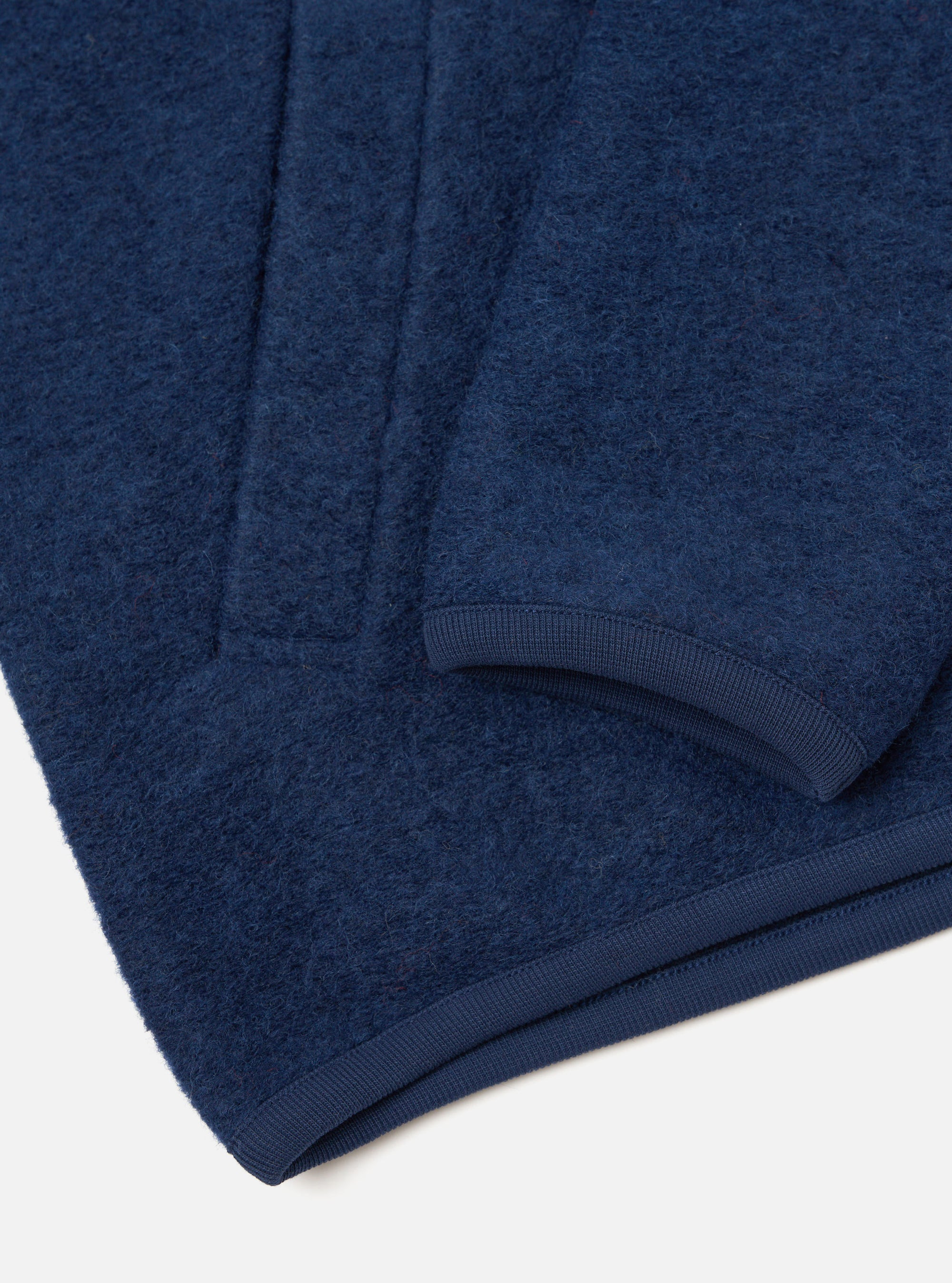 Close-up detail of an indigo wool fleece zip bomber, shot flat on a white surface. The brushed, textured fleece is visible in deep navy blue. The image focuses on the curved hem and short sleeve cuff, both finished with a smooth navy ribbed trim.