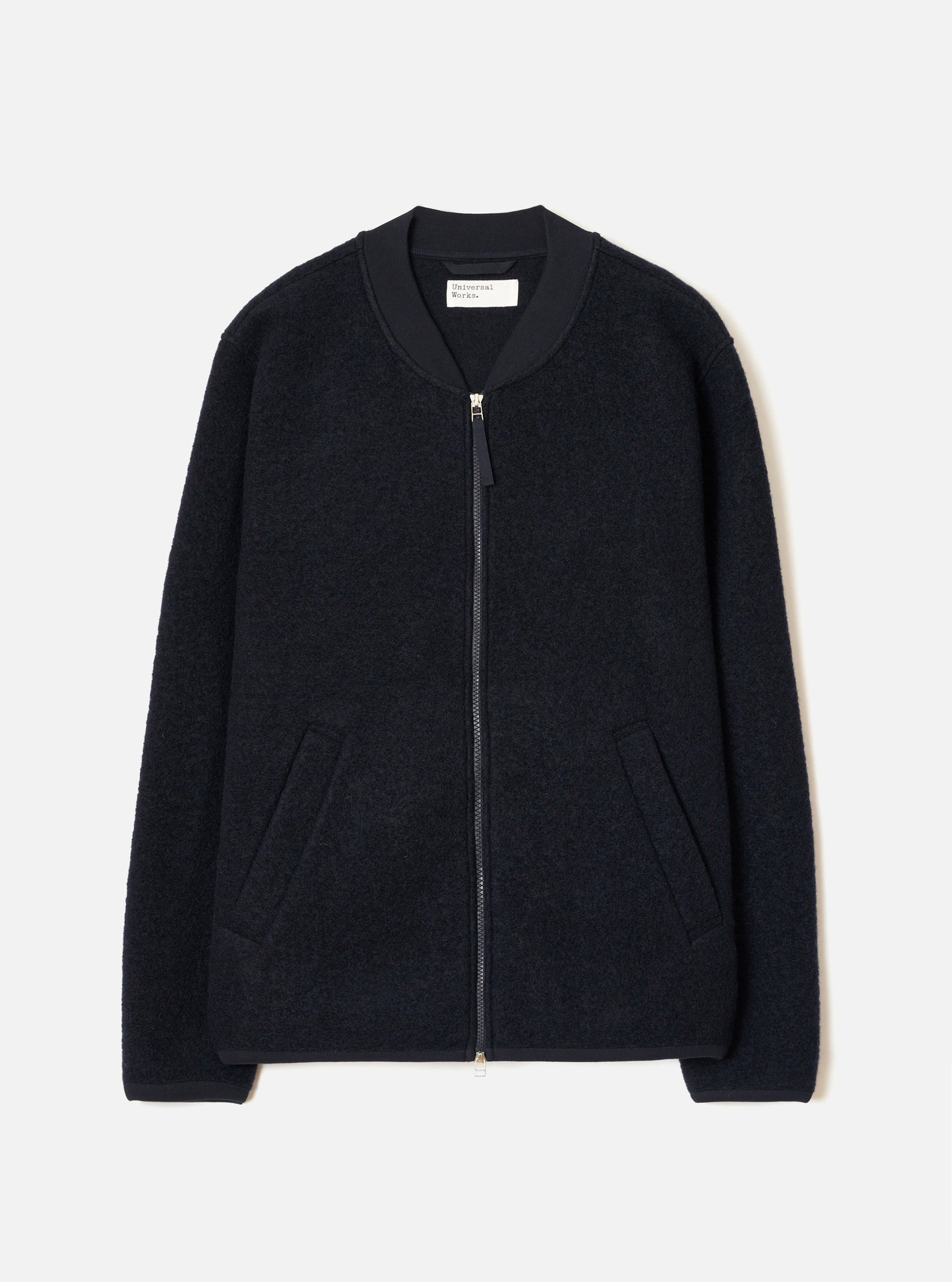 Navy wool fleece zip bomber jacket, flat lay on a light grey seamless backdrop. Textured boiled-wool-style fleece body with a smooth ribbed bomber collar in contrast navy fabric. Full-length silver zip with a small pull tab. Two welt pockets at the hip. Ribbed cuffs and hem. Universal Works woven label visible at the back neck.