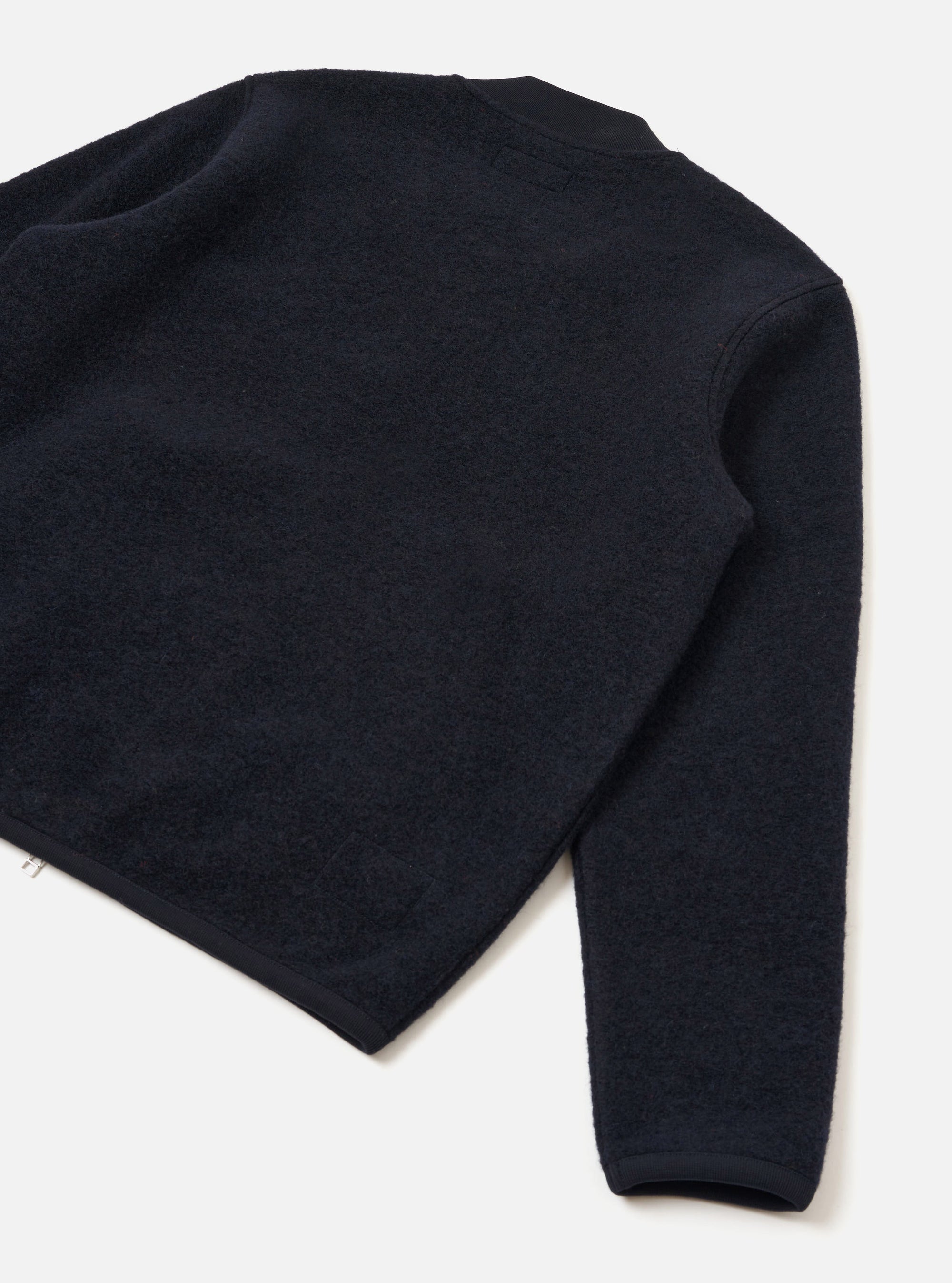 Navy wool fleece zip bomber laid flat on a white surface, shot from above at an angle showing the back and one sleeve. The textured fleece is trimmed with smooth navy grosgrain or woven binding at the crew neck, cuffs, and hem. A small silver zip pull is visible at the lower left hem.