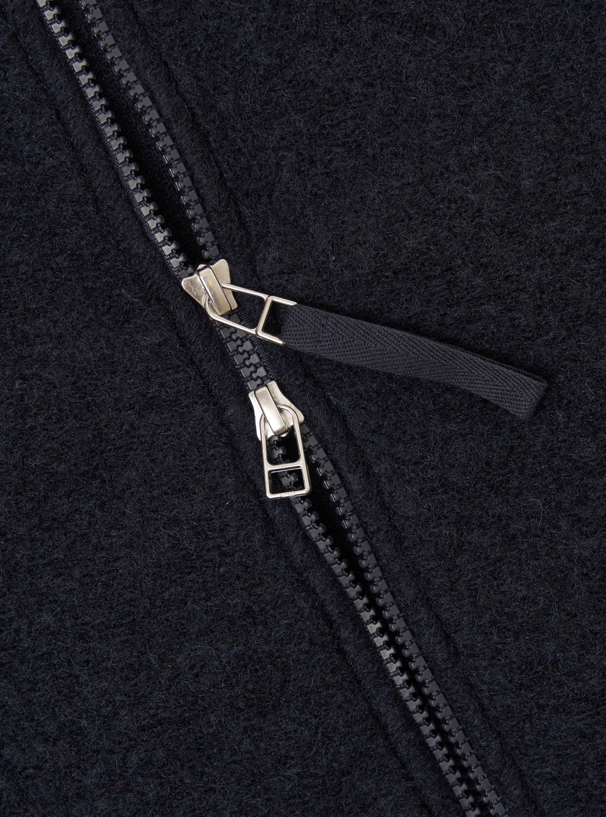Close-up detail of the double zip fastening on a navy wool fleece bomber jacket. Two silver-tone metal zip sliders sit centrally, with black plastic teeth and a dark woven zip pull tab. The brushed wool fleece fabric texture is visible across the frame.