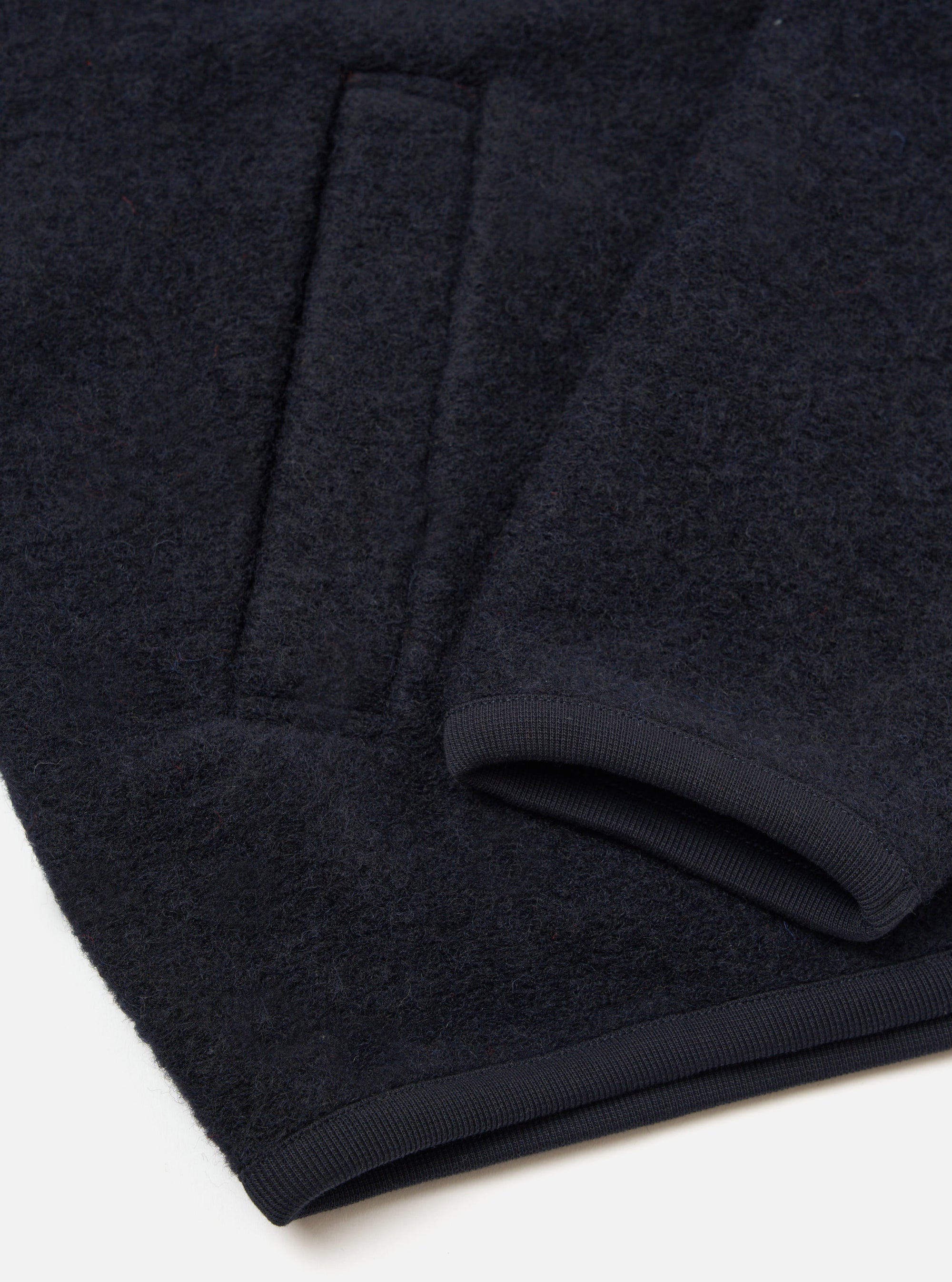 Close-up flat lay of a navy wool fleece zip bomber on a white surface. The boiled wool texture is clearly visible. The shot shows the lower front panel, a chest welt pocket, the ribbed hem, and a ribbed cuff folded back to reveal its trim detail.
