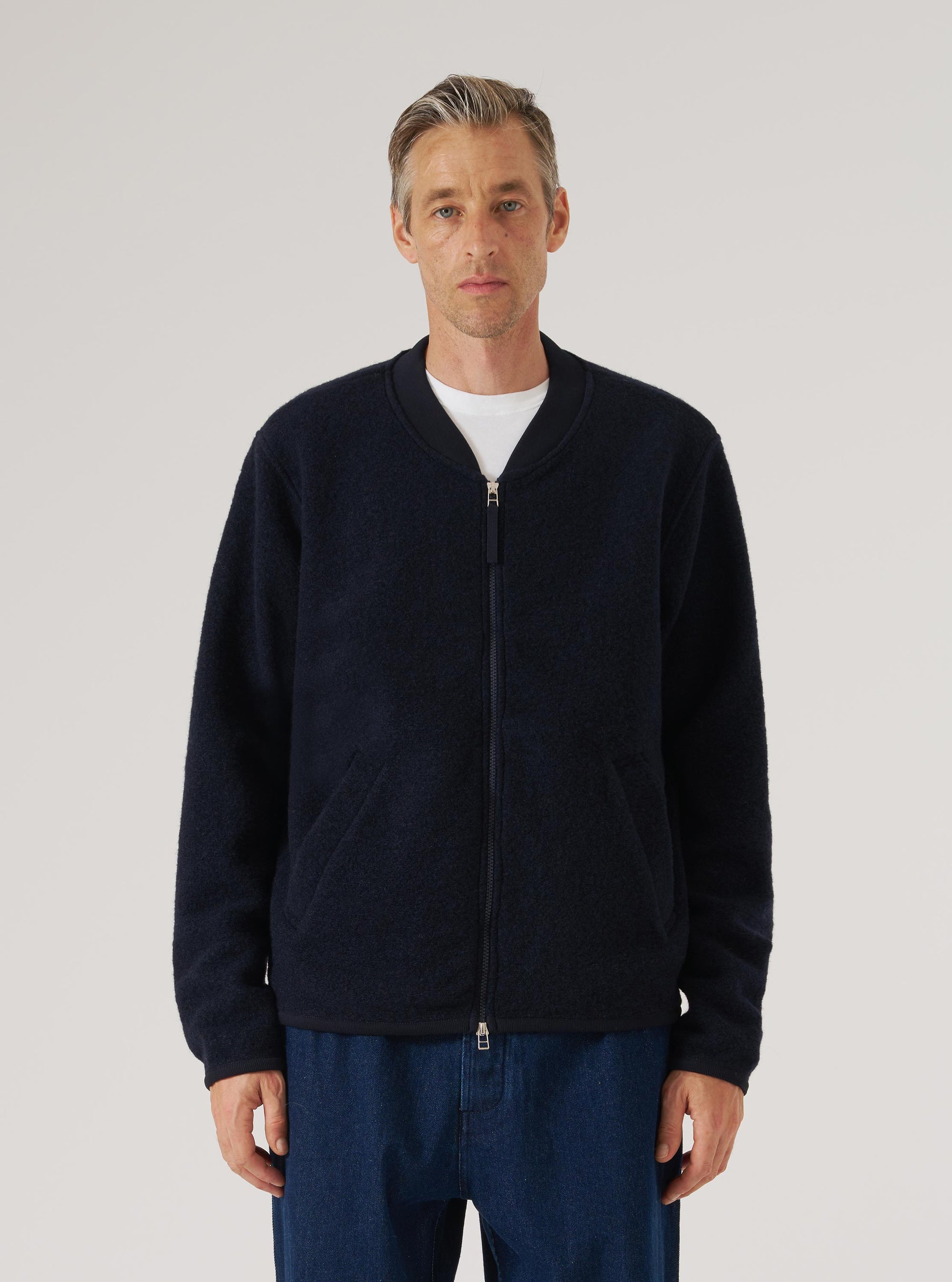 Navy wool fleece zip bomber worn by a male model against a light grey studio backdrop. The jacket is fully zipped, with a silver two-way zip, ribbed bomber collar, and two slanted side pockets. The textured boiled wool fleece fabric is visible throughout. Cropped at the thigh, layered over a white crew-neck tee and dark blue jeans.