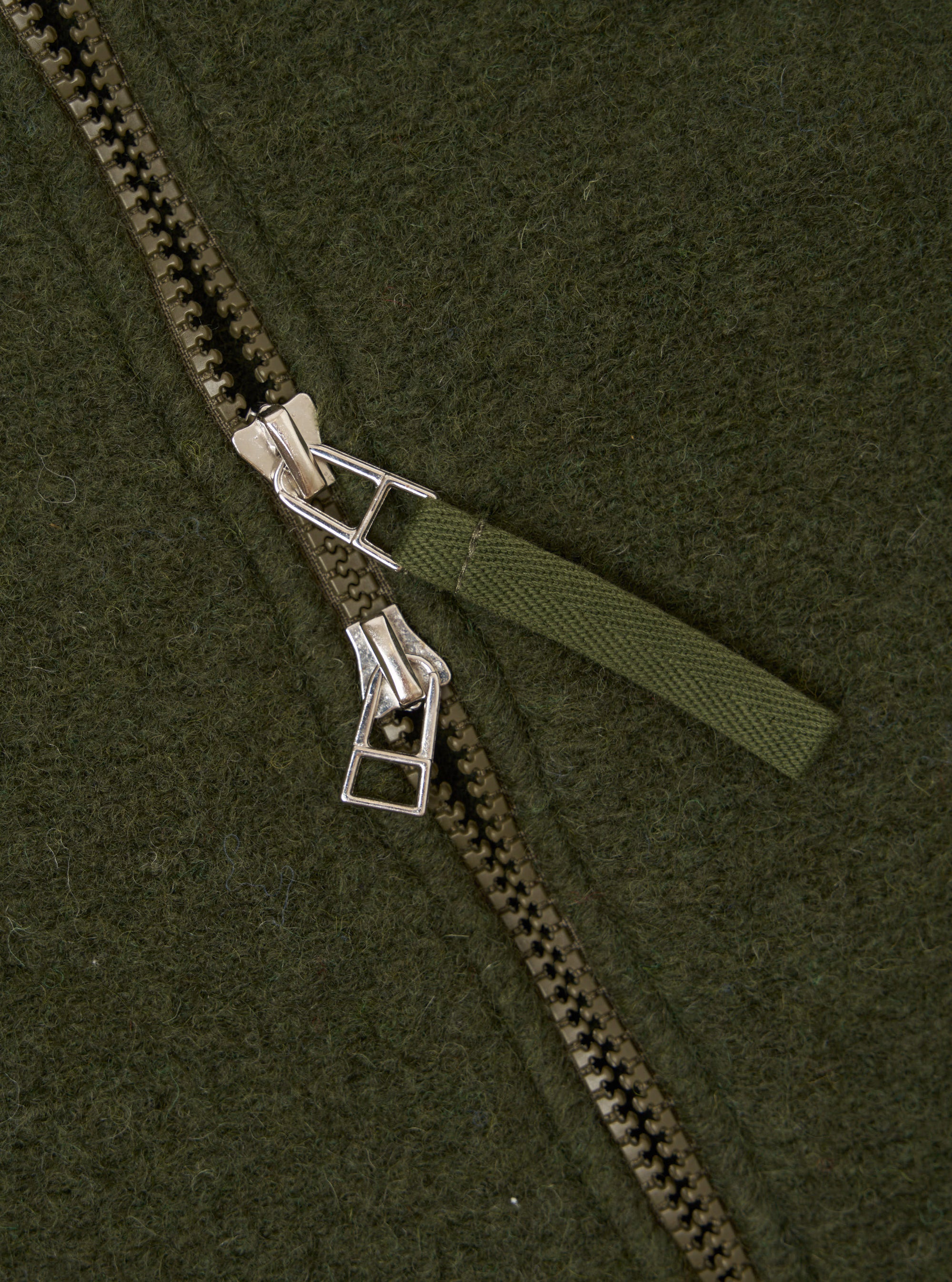 Close-up detail of the zip on an olive wool fleece bomber jacket. A chunky two-way zip with silver metal sliders and an olive woven fabric pull tab runs diagonally across the frame. The brushed wool fleece texture is visible across the background.