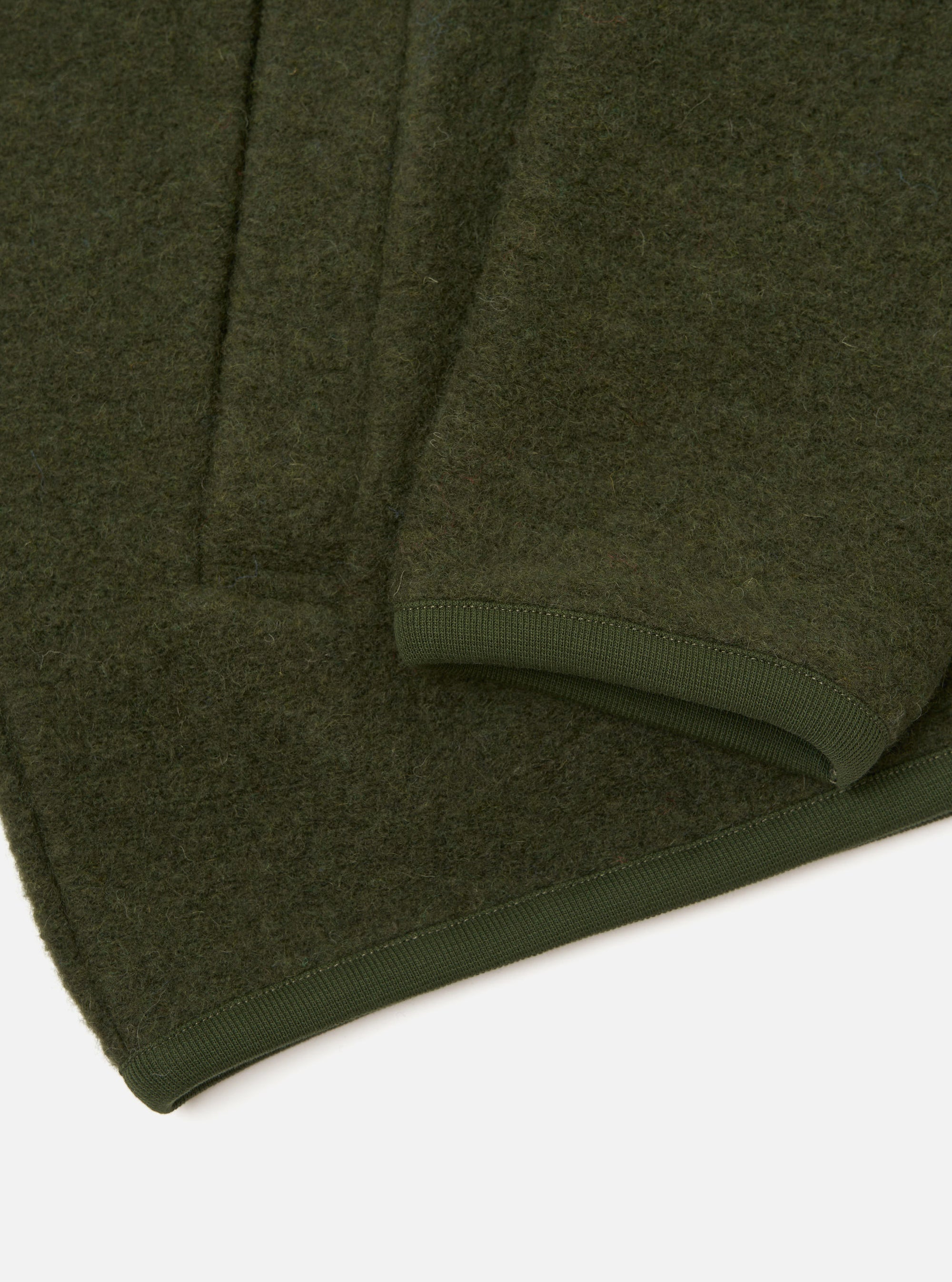 Close-up flat lay of an olive wool fleece zip bomber on a white surface. The boiled wool texture is visible across the body. A ribbed knit hem trim in matching olive runs along the bottom edge, with neat topstitching. A sleeve fold is visible in the upper portion of the frame.