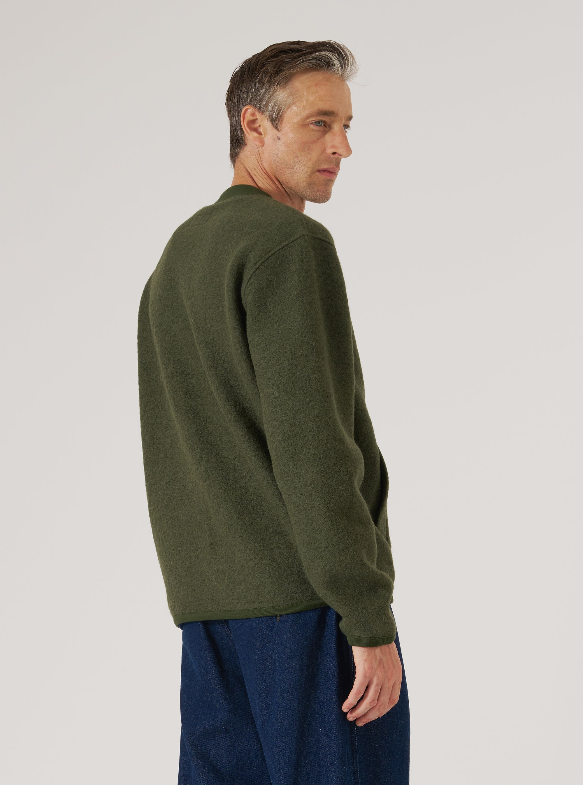 Male model photographed from behind and to the side against a light grey studio backdrop, wearing an olive wool fleece zip bomber with a ribbed crew neck collar and long sleeves with ribbed cuffs. The brushed wool texture is visible across the back. Worn untucked over dark navy trousers. Shot crops just below the hips.