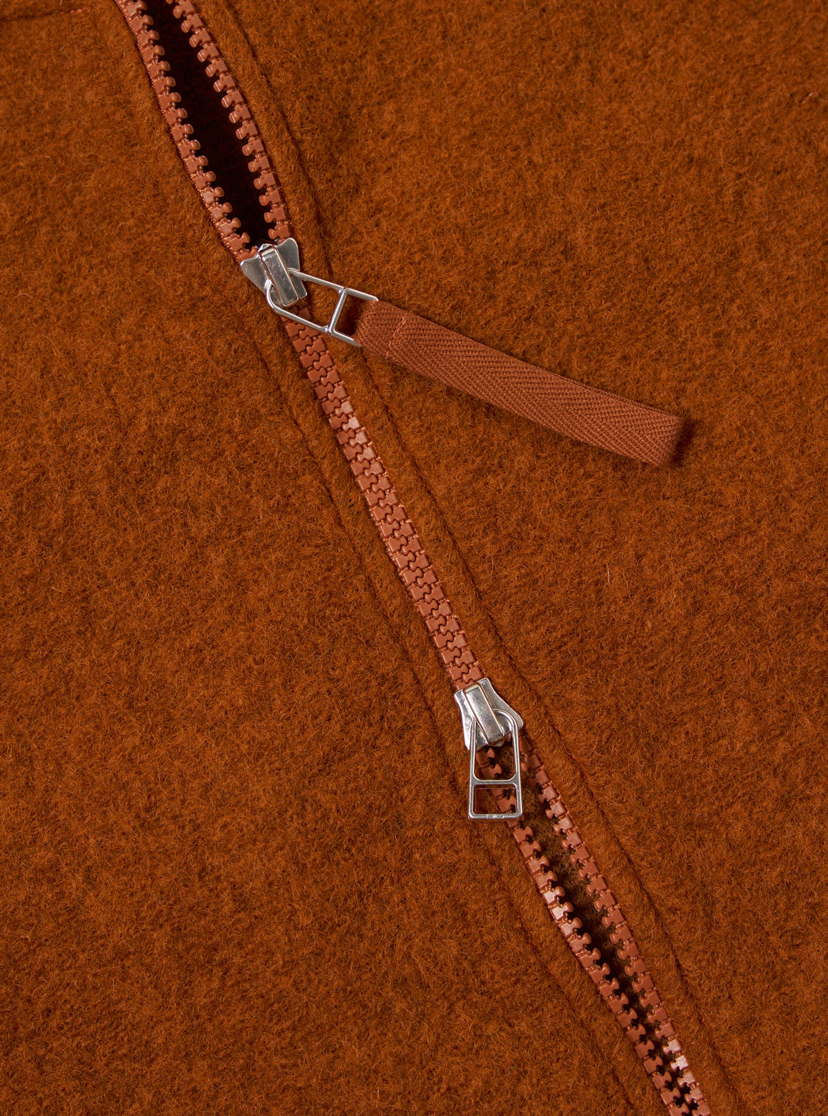 Close-up of the rust-colored Wool Fleece Zip Bomber, showcasing the texture and zipper detail. The background is a simple, clean surface.