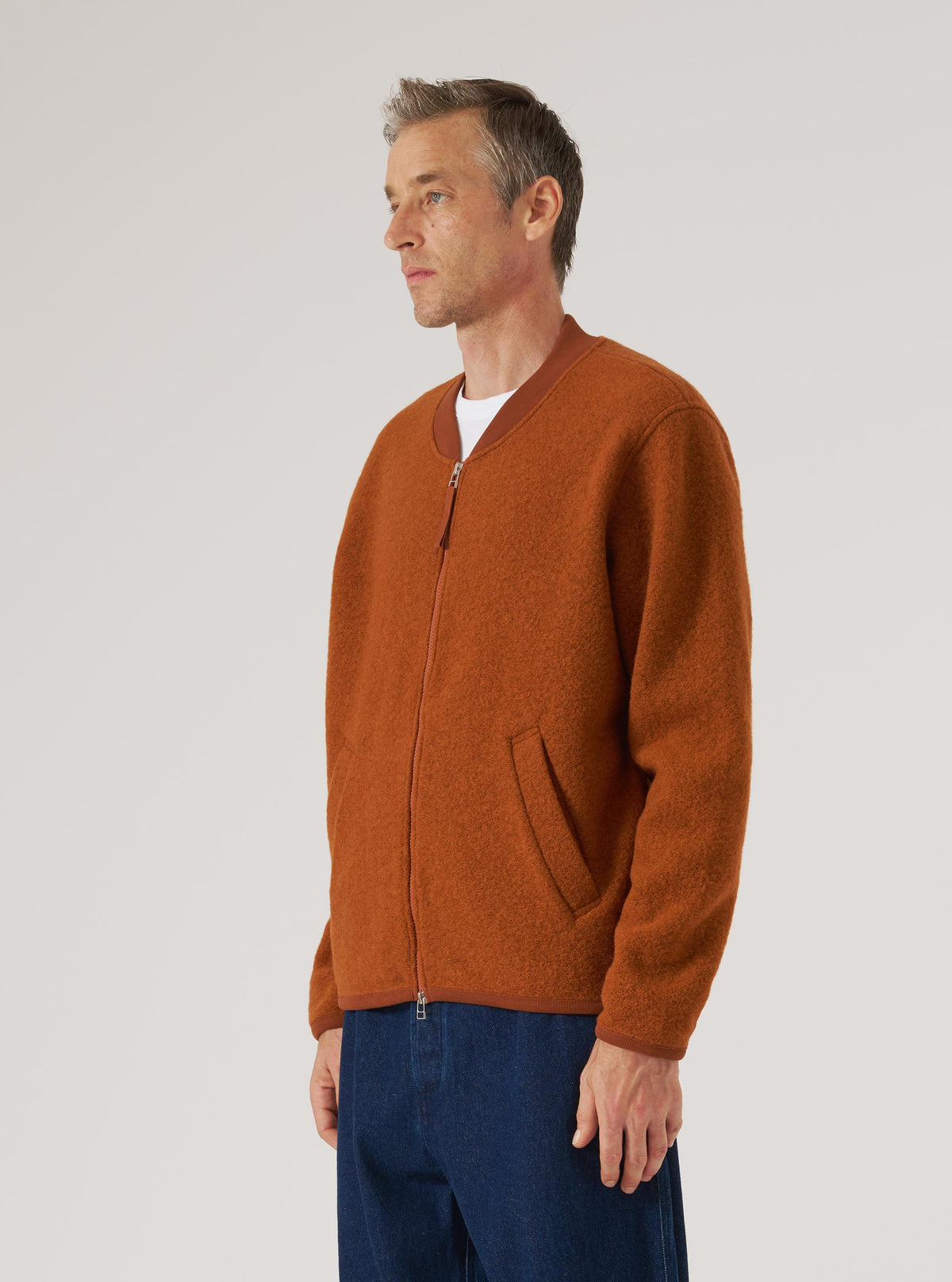 Profile of a man wearing a Rust Wool Fleece Zip Bomber in rust color. The background is a plain white. The bomber has a zip closure and ribbed trim.