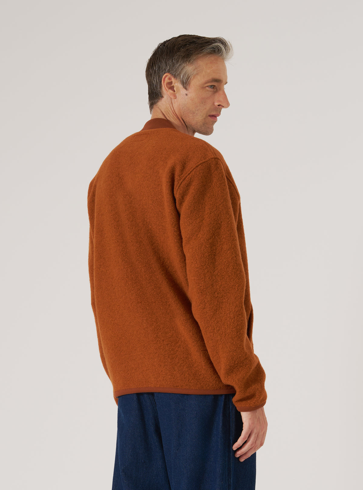 Profile of a man in a rust-colored wool fleece zip bomber jacket against a white backdrop. The jacket has a relaxed fit and is paired with dark wash jeans.
