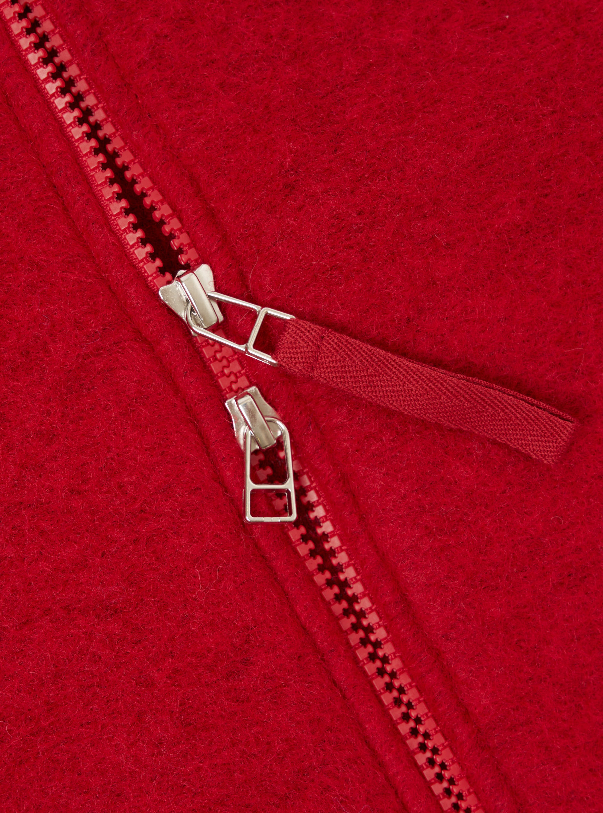 Close-up detail of the zip on a scarlet wool fleece bomber. A two-way zip with red plastic teeth and two silver metal sliders is shown partially open. A red woven zip puller tab is attached to the upper slider. The brushed wool fleece fabric texture is visible across the background.