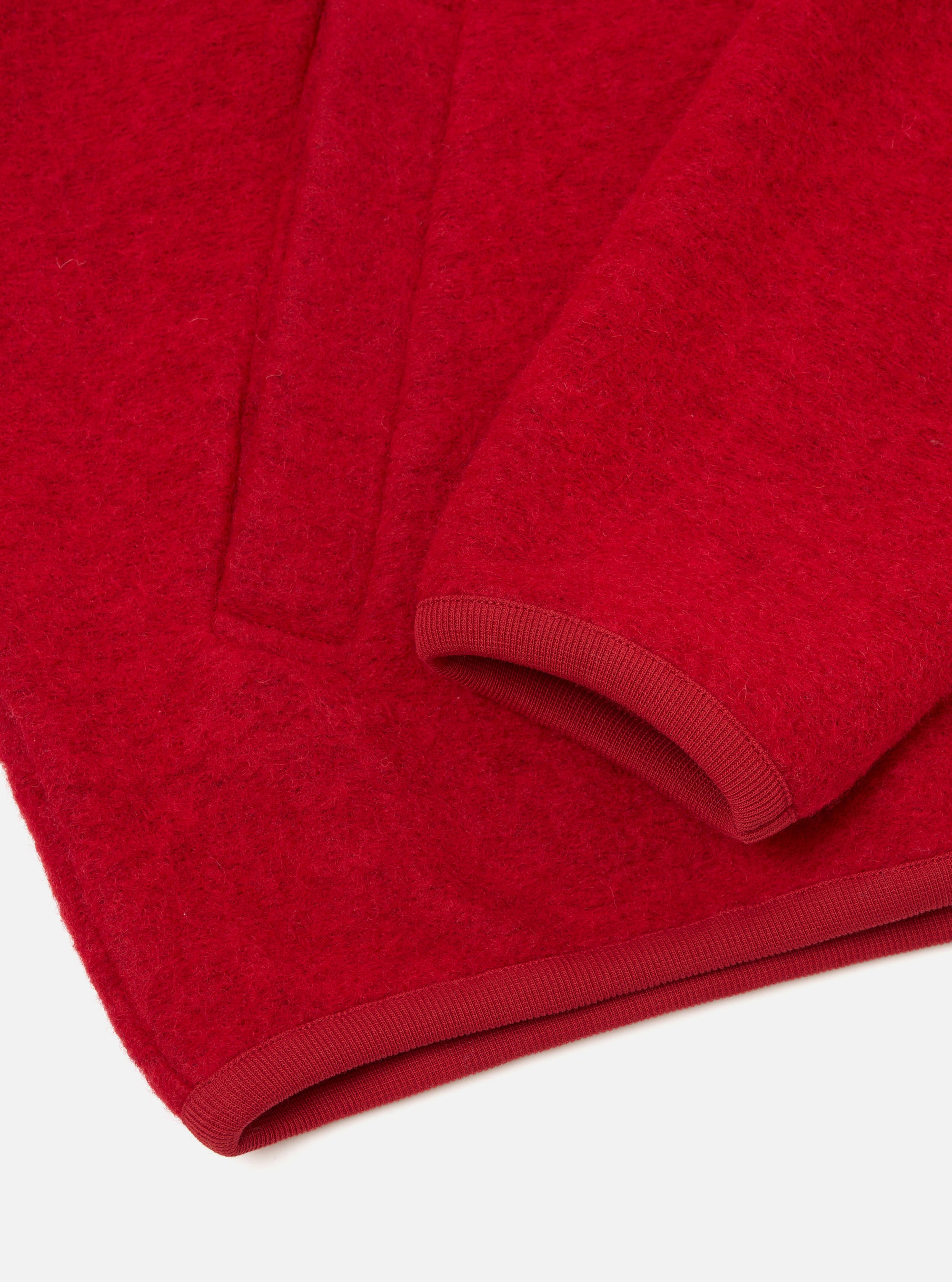 Close-up flat lay of a scarlet wool fleece zip bomber on a white surface. The boiled wool texture is clearly visible. A ribbed knit cuff curves at the sleeve opening, with a matching ribbed hem band running along the bottom edge.