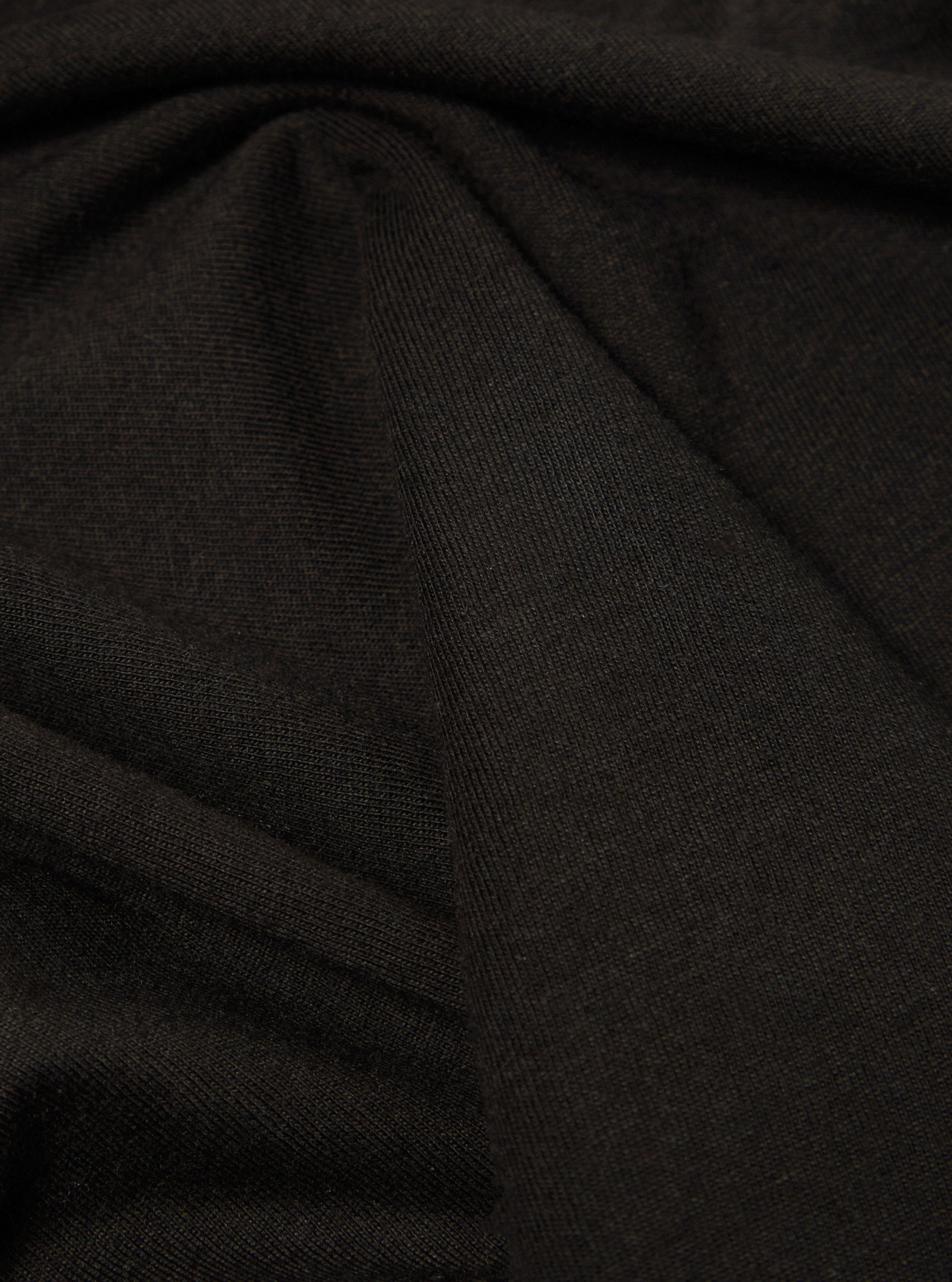 Close-up of the Black Single Jersey Core Tee fabric. The soft, single jersey material is draped to show texture. Dark, neutral background.