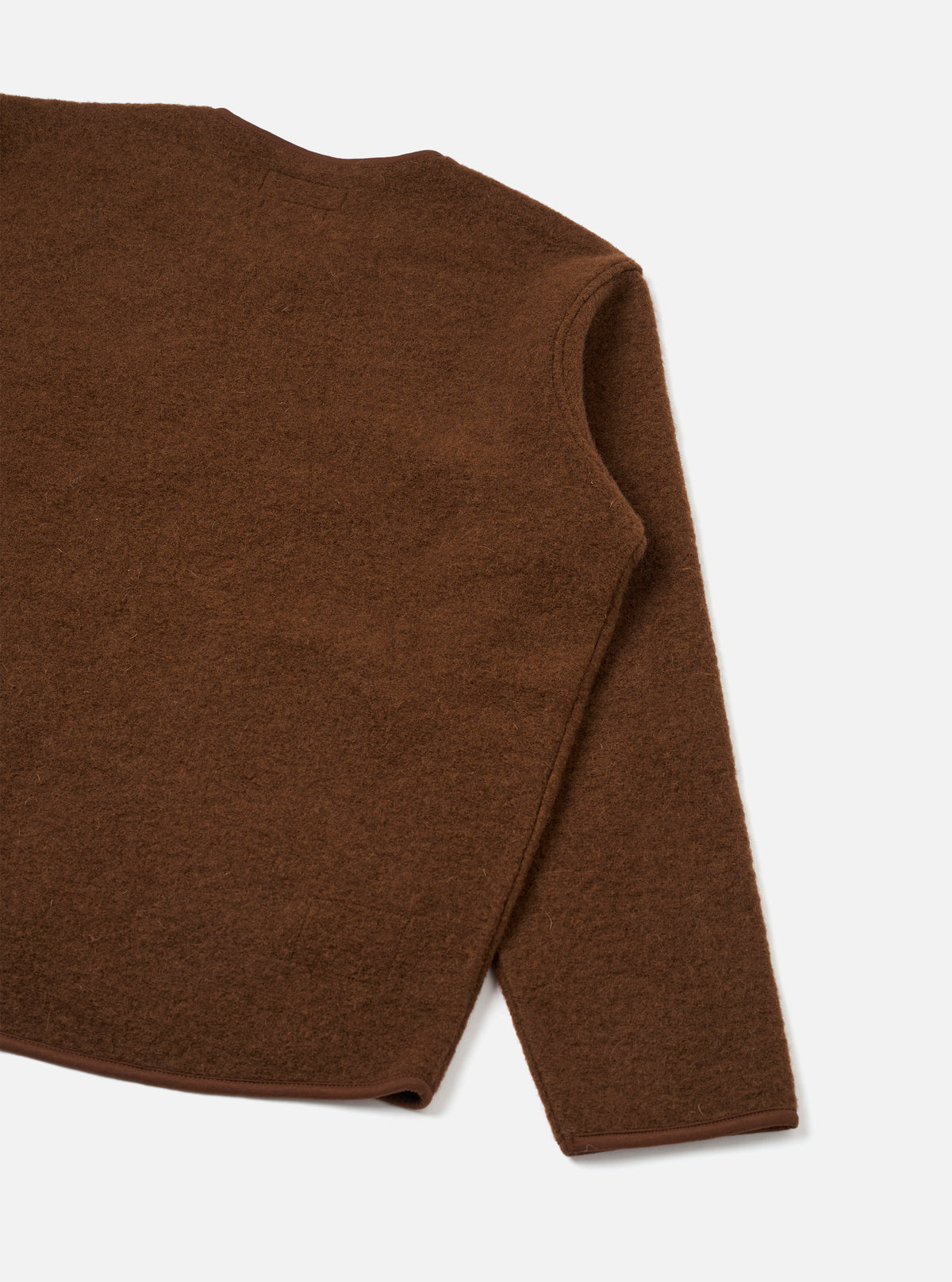 Close-up flat lay of a cacao brown wool fleece zip liner jacket on a light grey seamless backdrop. The shot captures the upper back, right shoulder seam and one full sleeve. The boiled wool fleece has a lightly textured, brushed surface. A narrow bound hem edge is visible at the sleeve and bottom.