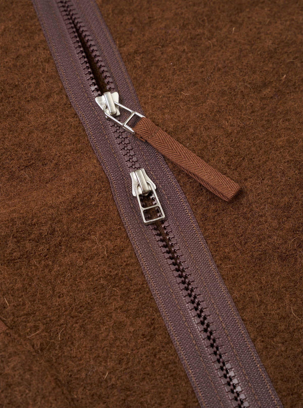 Close-up detail of the double zip fastening on a cacao wool fleece liner jacket. Two silver-hardware zip sliders sit on a dark plum zip tape with moulded plastic teeth. A woven brown zip garage tab is visible. The brushed, textured wool fleece fabric fills the background.