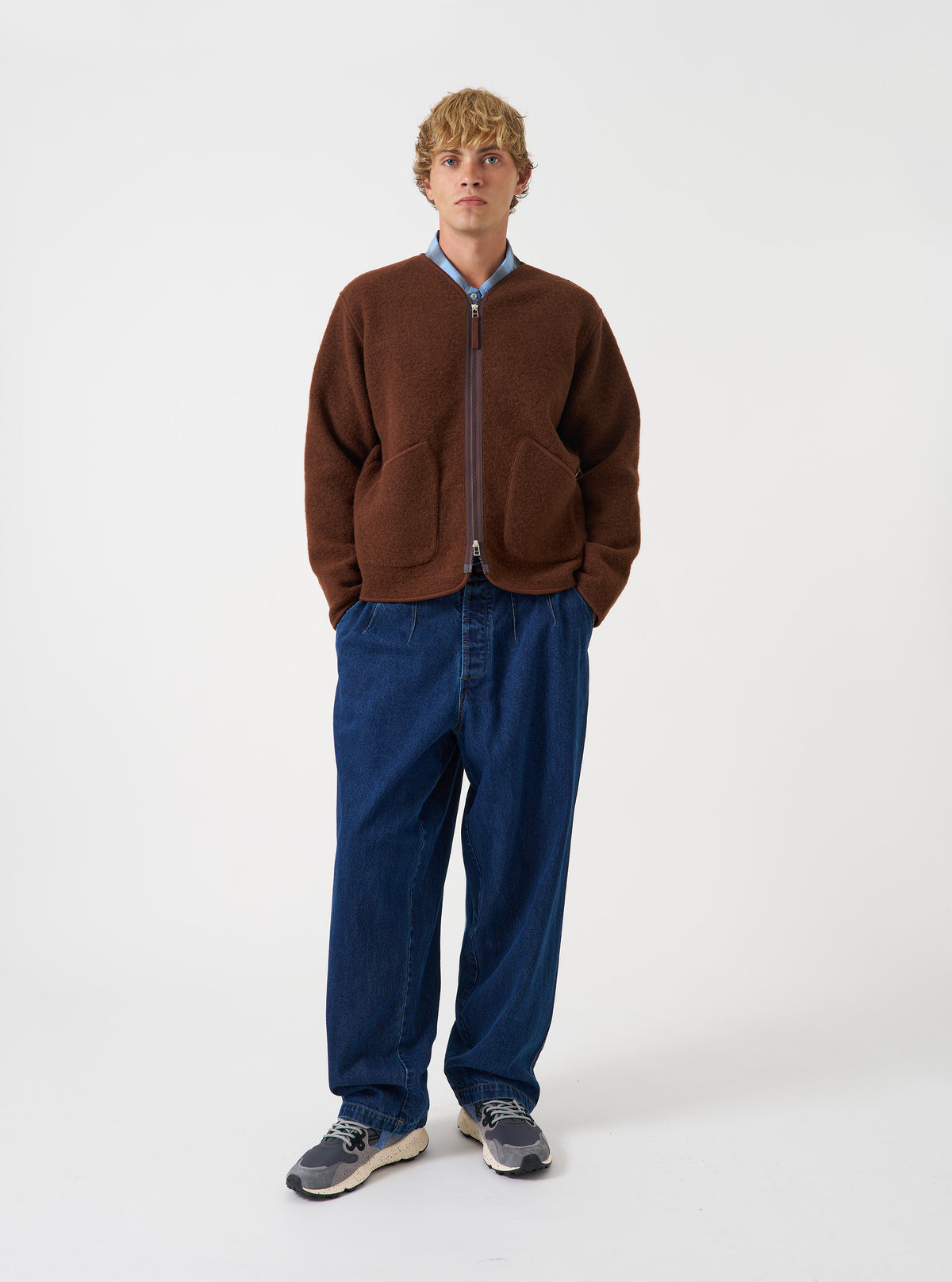 Front view of a model wearing the Cacao Wool Fleece Zip Liner Jacket in a studio setting. The model stands facing forward, displaying the brown wool fleece material, central front zip, collarless neck, and relaxed layer fit.