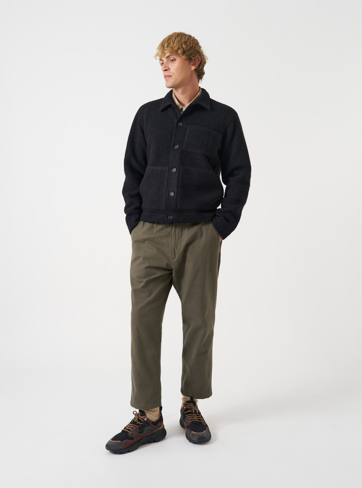 Front view of a model wearing the Black Wool Fleece Uniform Jacket in a brightly lit studio with a plain backdrop. The model stands facing forward, showing the classic collar, front button closure, and chest pockets.