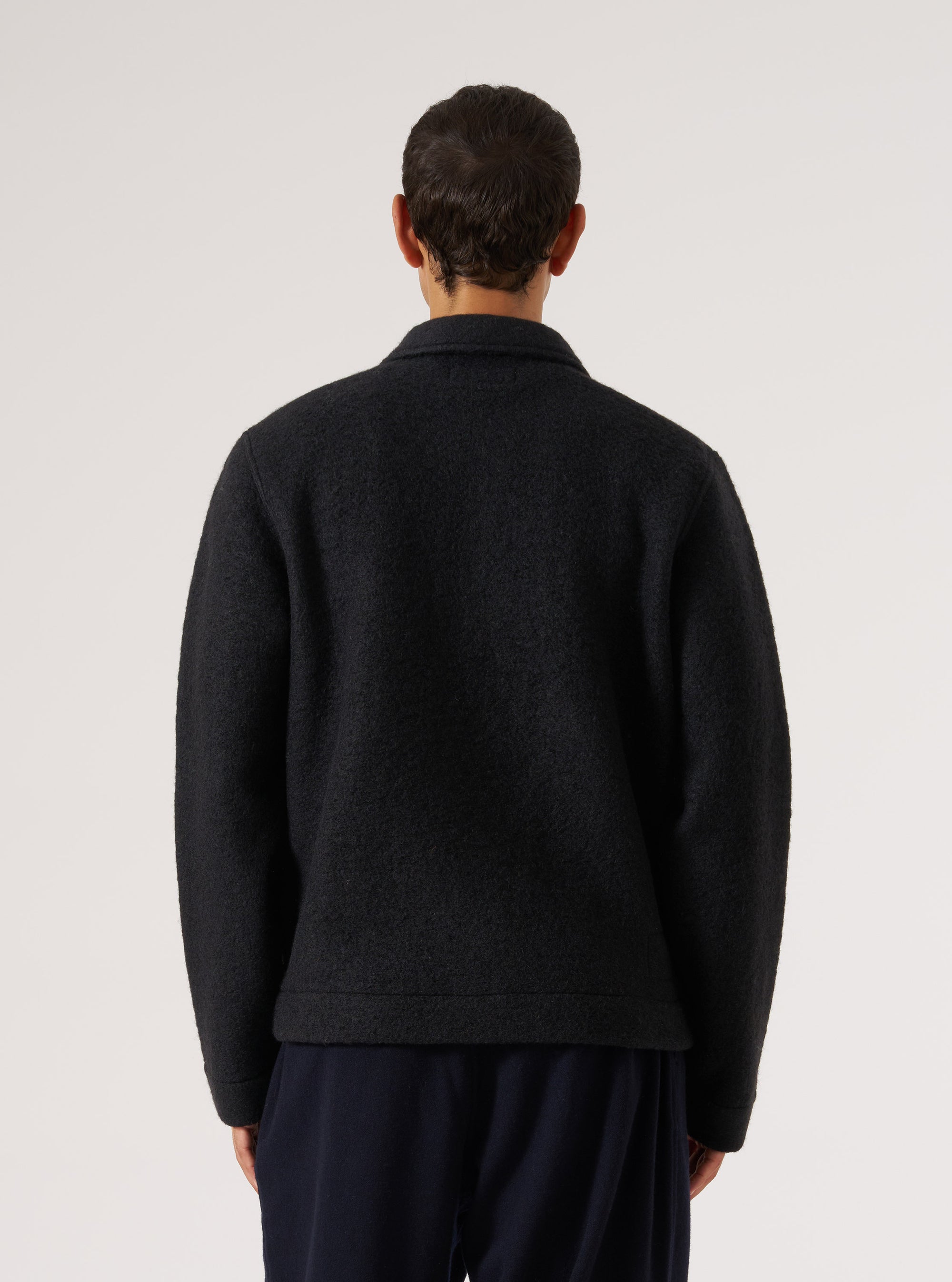 Back view of a male model wearing a black wool fleece uniform jacket against a light grey studio backdrop. The textured fleece fabric is visible across the back panel and full-length sleeves. A spread collar is visible at the neckline. The jacket has a ribbed hem and relaxed fit. Worn over navy trousers. Shot cropped from mid-thigh up.