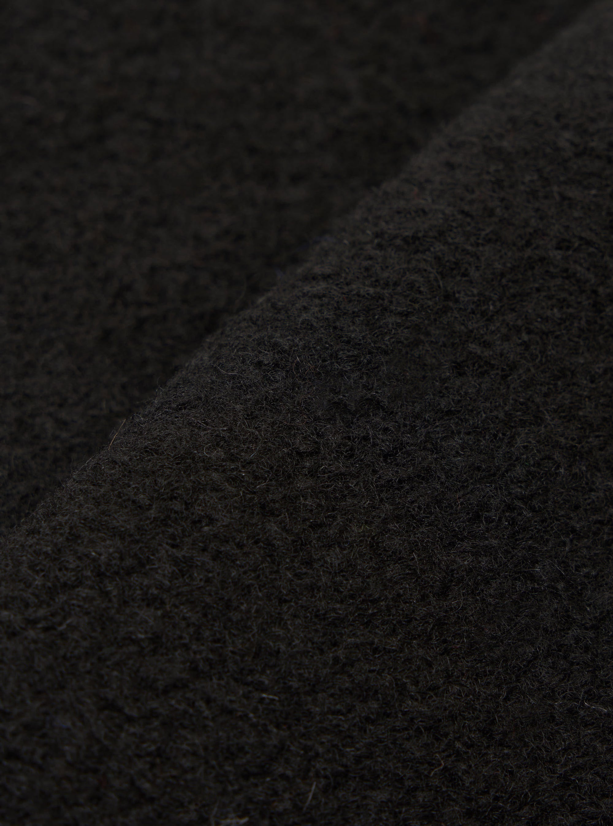 Close-up fabric detail of the black wool fleece uniform jacket, showing the dense, nubby texture with fine looped fibres across the surface. A folded seam or edge runs diagonally through the frame, highlighting the fabric's depth and weight.