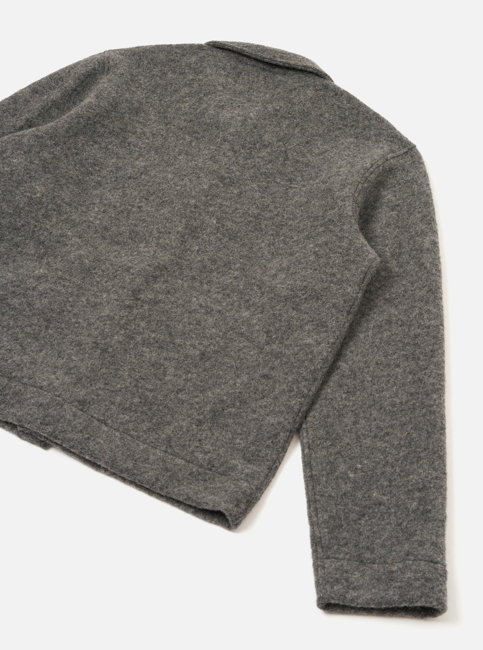Close-up flat lay of a grey marl wool fleece uniform jacket on a white studio surface, shot from above at an angle. The back panel and one sleeve are visible, showing the soft, lightly textured boiled wool fleece fabric. A small collar is visible at the top. The sleeve ends in a plain, clean-finished cuff.