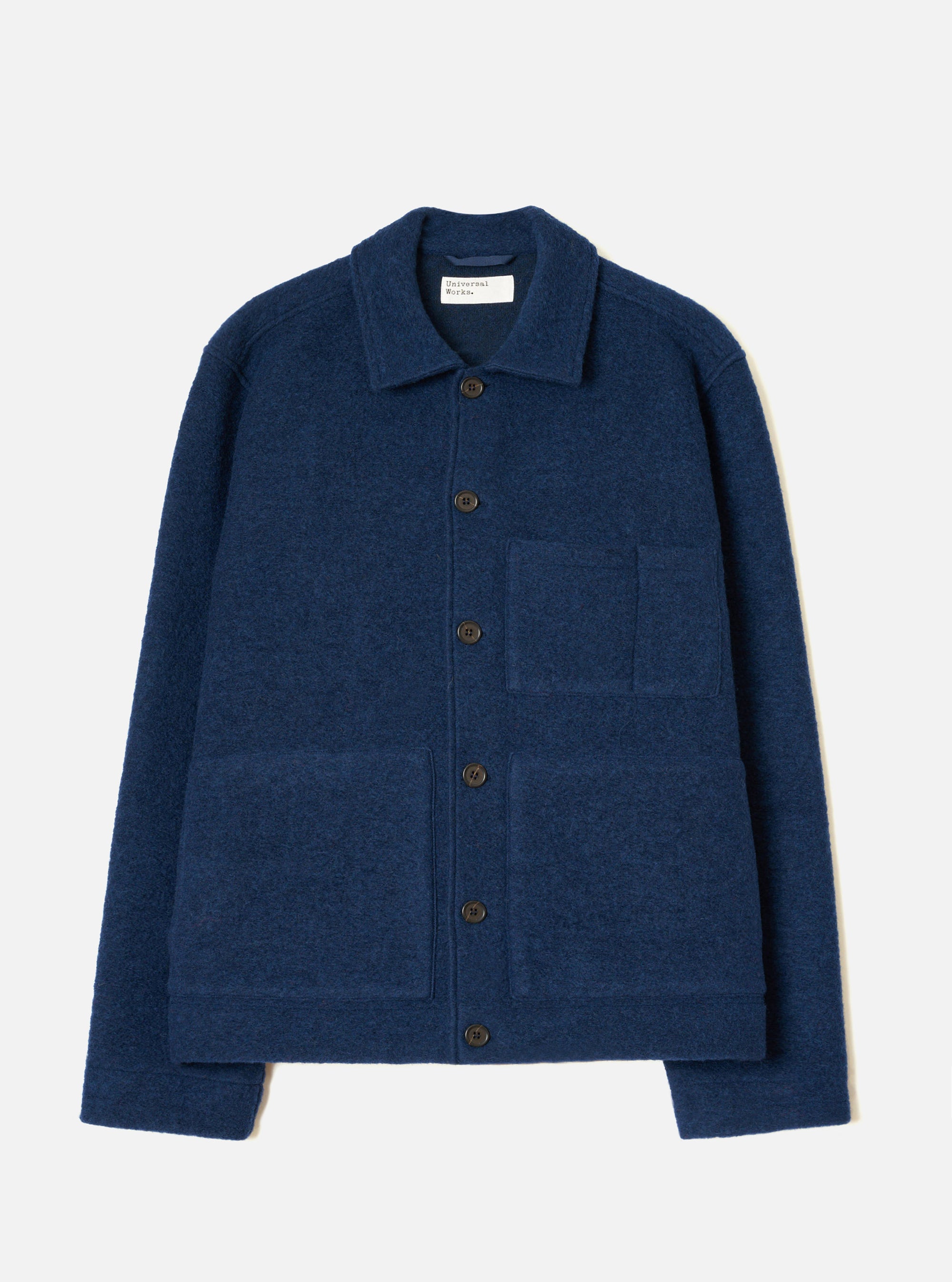 Indigo wool fleece uniform jacket laid flat on a light grey studio backdrop. Features a spread collar, five dark horn-effect buttons down the centre front, one chest patch pocket, two large lower patch pockets, ribbed cuffs, and a ribbed hem. The textured boiled wool fleece fabric is visible throughout.