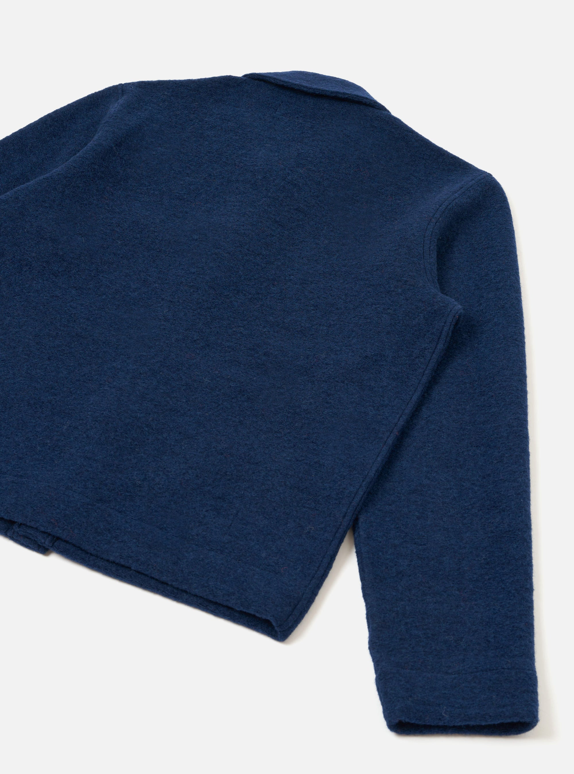 Rear three-quarter flat lay of an indigo wool fleece uniform jacket on a light grey seamless backdrop. The boiled wool texture is clearly visible across the back panel. A small collar is partially visible at the top. The right sleeve extends to a plain, unfinished cuff. The cropped hem sits straight and clean.